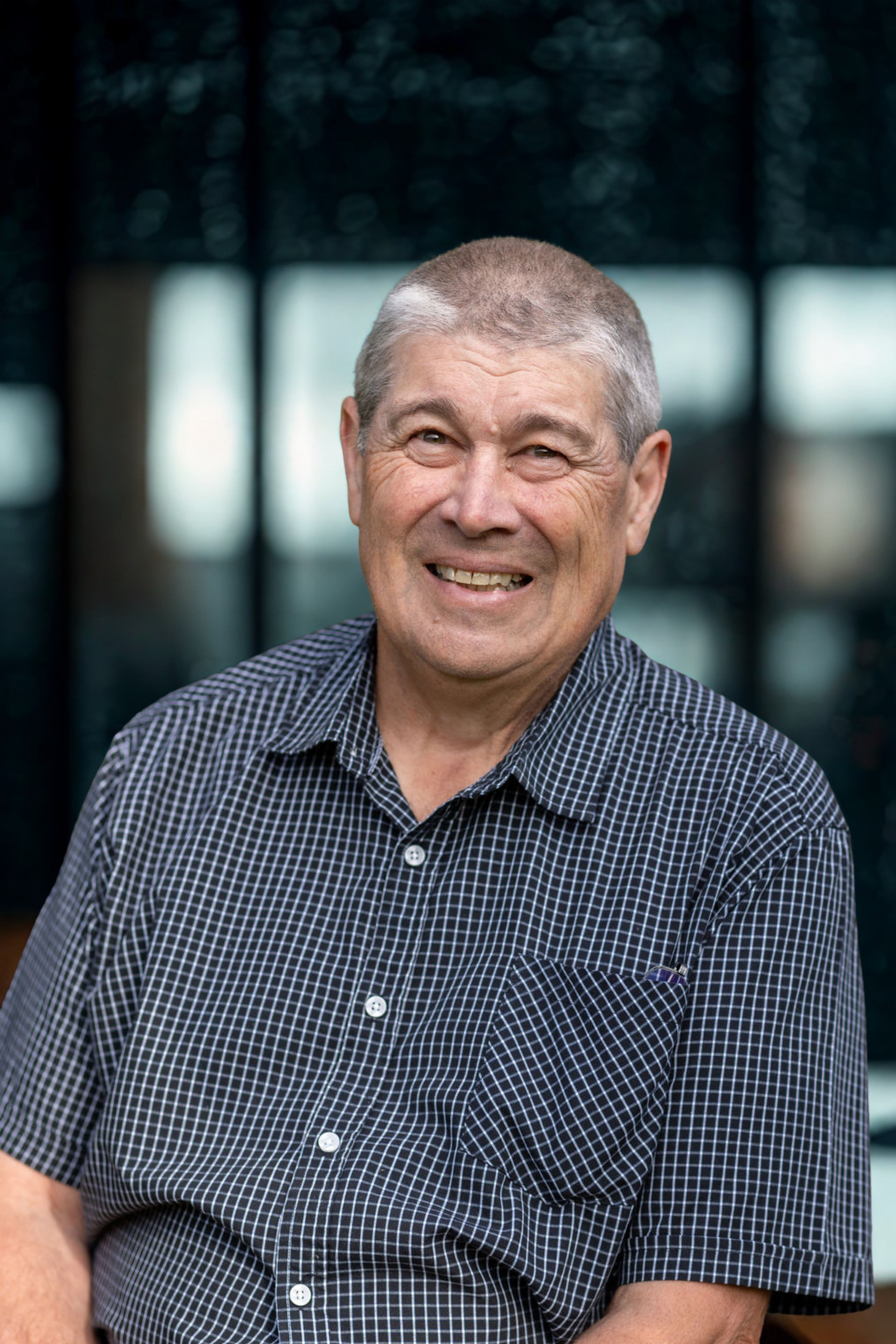 Close-up of a smiling middle-aged man with short gray hair, wearing a black and white checkered shirt, standing outdoors with a blurred background.