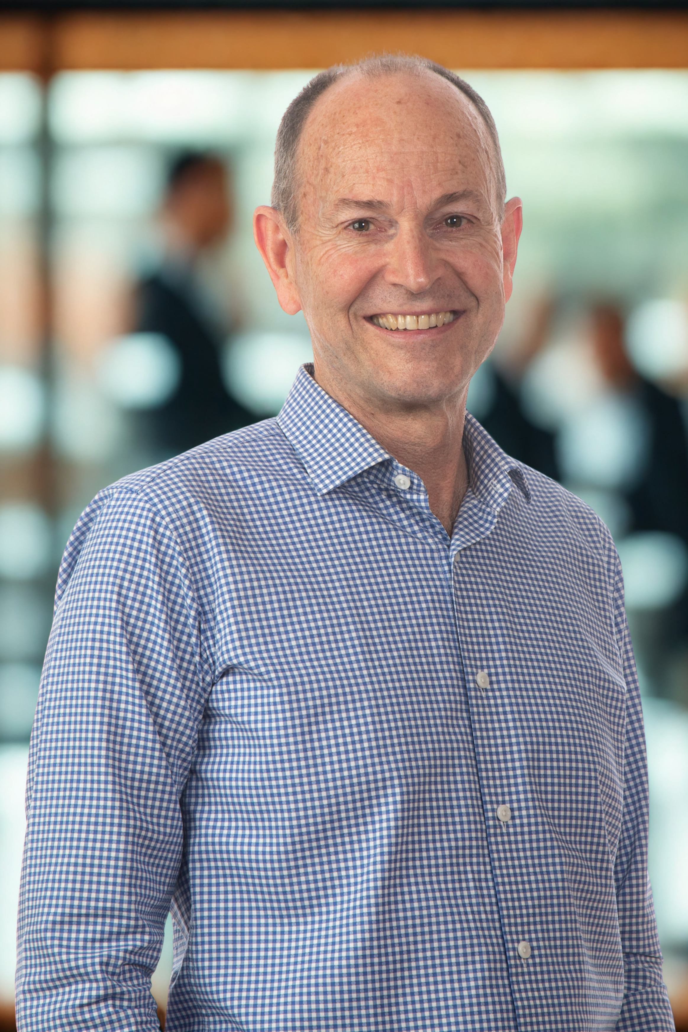 Smiling middle-aged man with short hair and a checkered shirt standing indoors, blurred figures in the background.