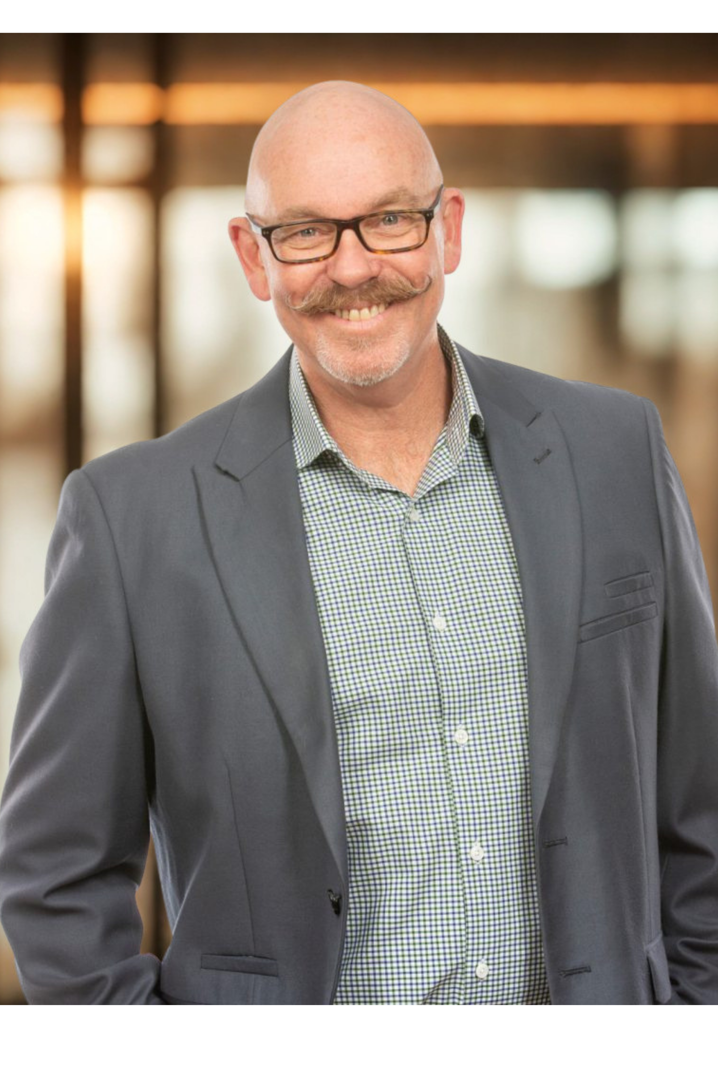 A bald man with glasses and a styled mustache, smiling, wearing a gray suit and a checkered shirt, standing indoors with blurred warm lighting in the background.