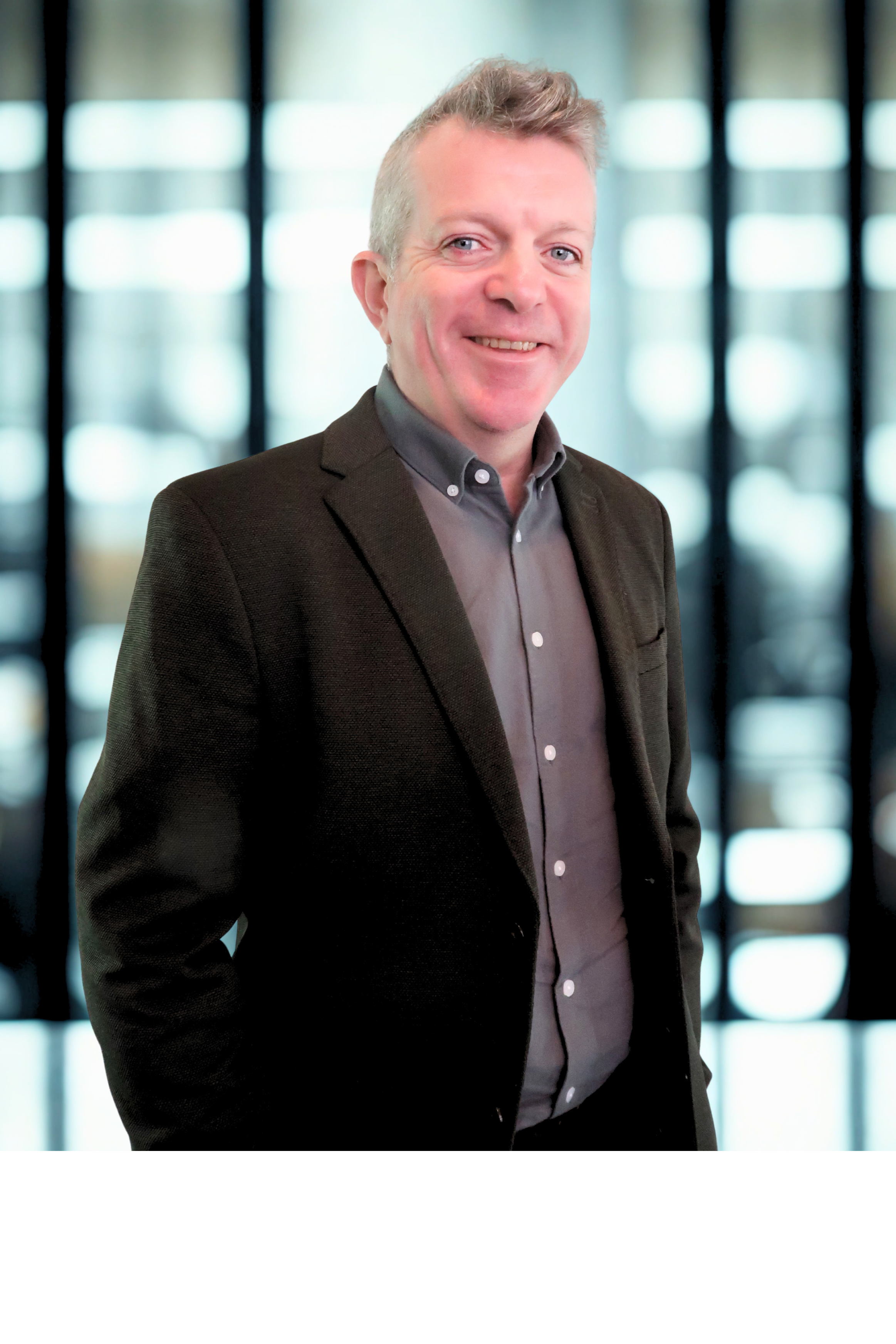 A professional man in a dark blazer and gray shirt standing in front of a blurred background of illuminated windows.