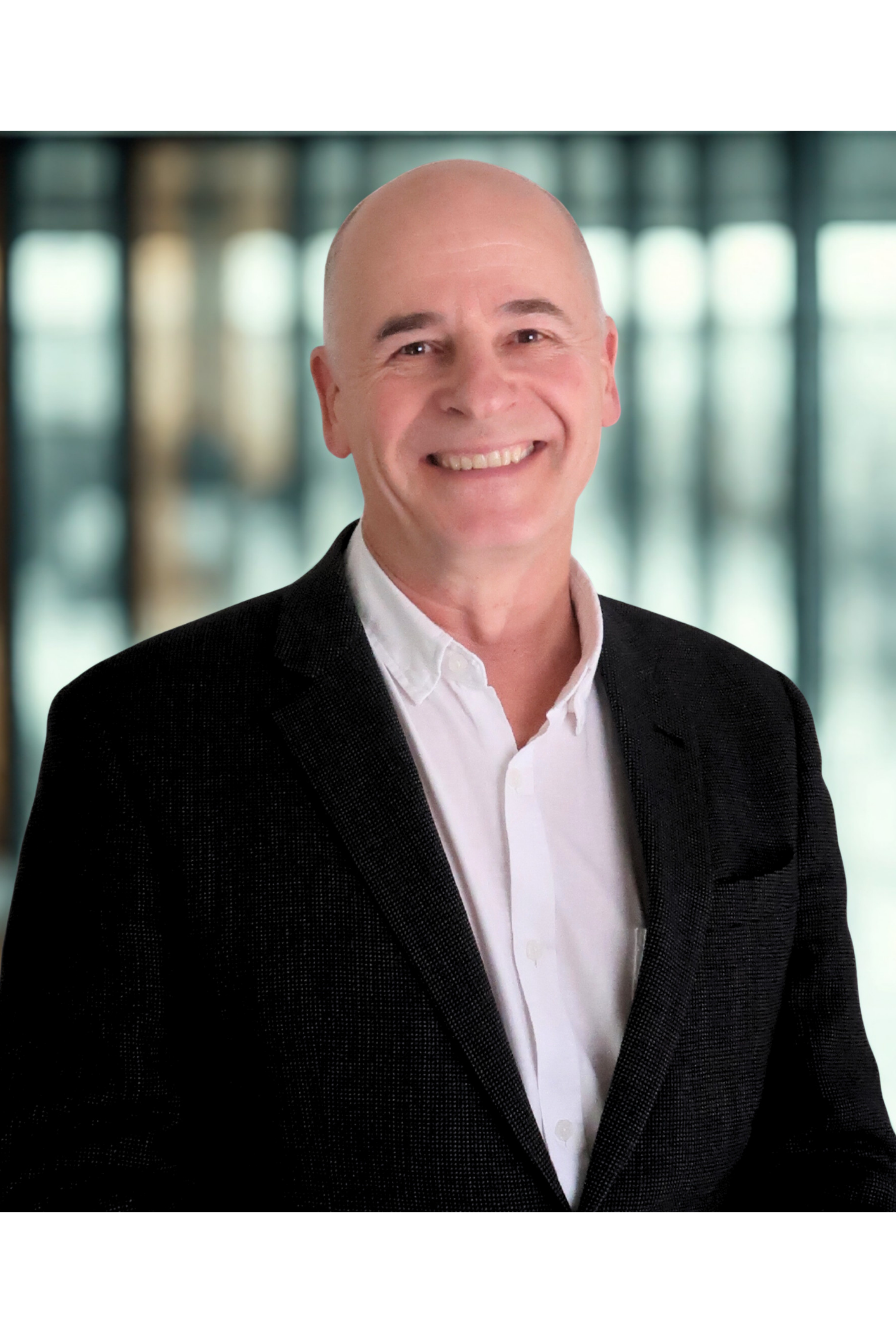Headshot of a smiling, bald man in a black suit jacket and white shirt, against a blurred modern office background with large windows.