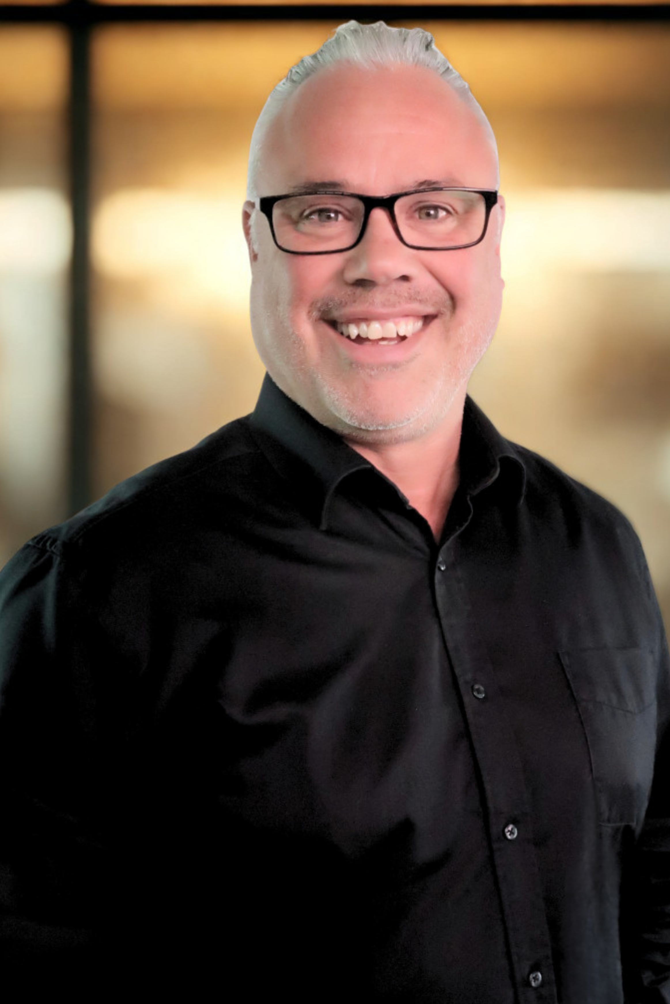 Portrait of a smiling man wearing black glasses and a black shirt, background is blurred with warm lighting.