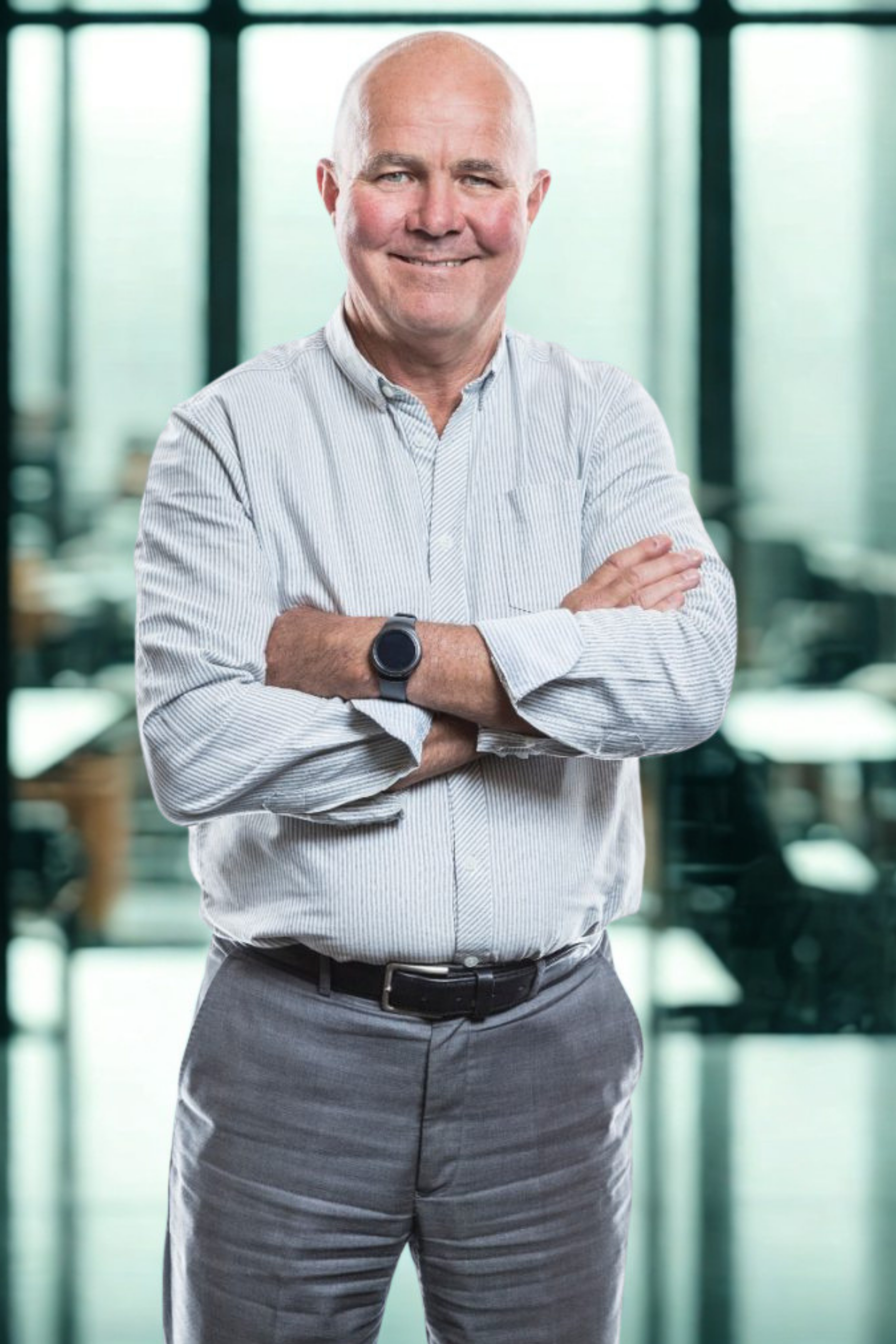 A smiling bald man with crossed arms, wearing a light striped shirt and gray pants, standing in an office setting with large windows in the background.