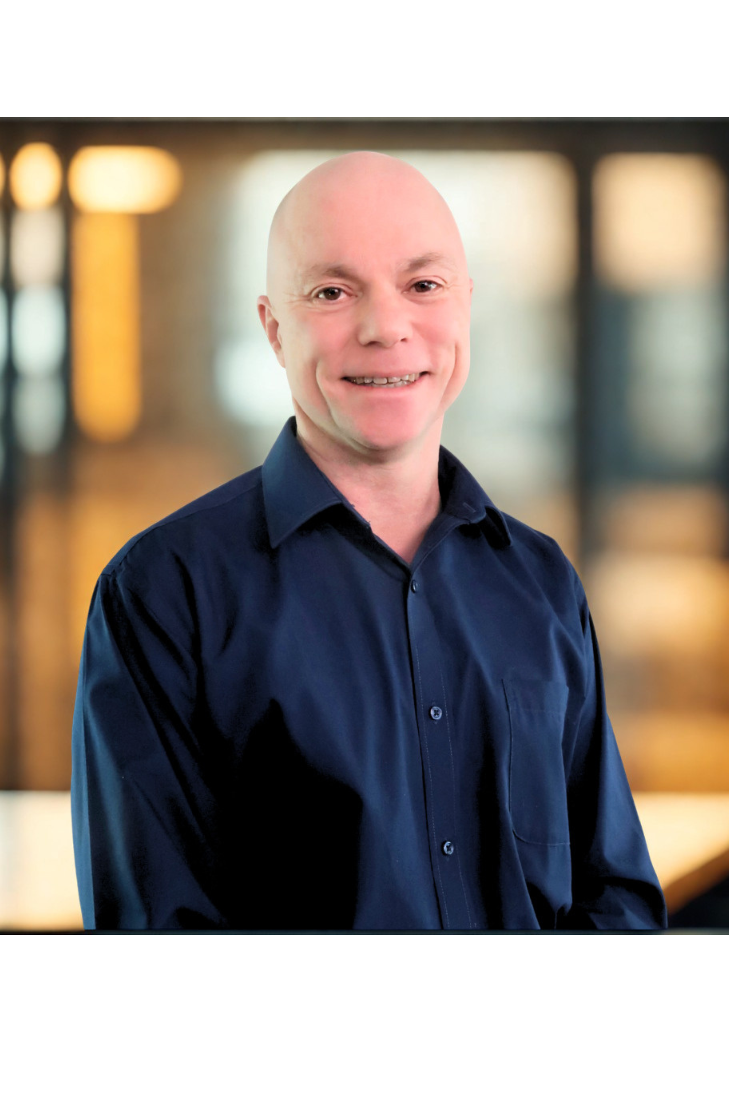 A man with a shaved head and a friendly smile, wearing a navy blue button-up shirt, standing indoors with blurred warm lighting in the background.