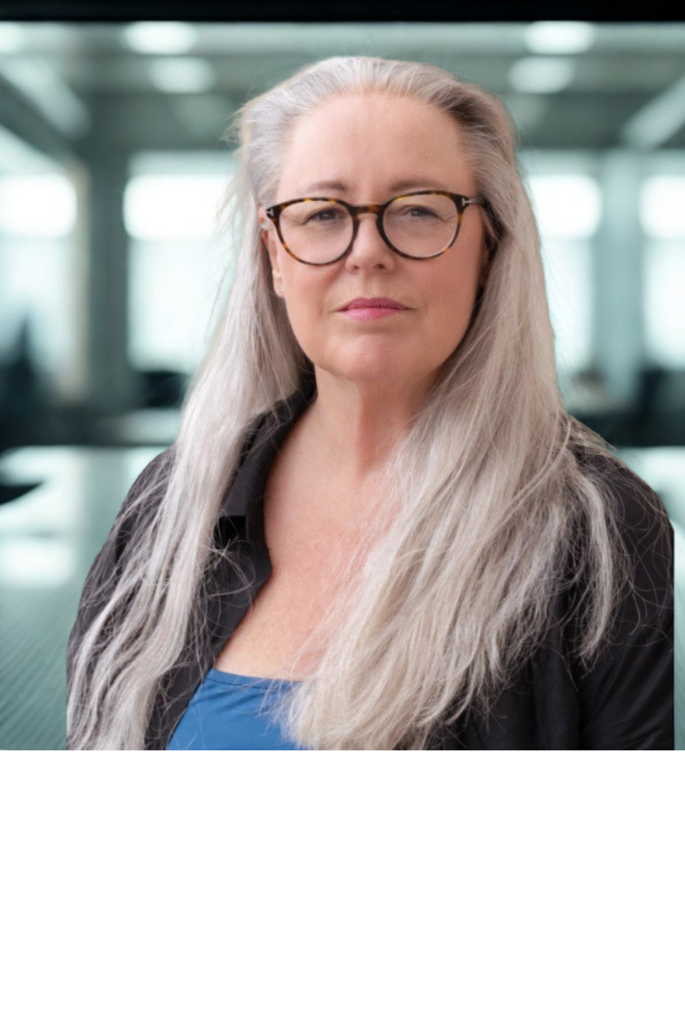 A middle-aged woman with long gray hair, wearing glasses, a black jacket, and a blue top, stands in an indoor space with large windows and a blurred background.