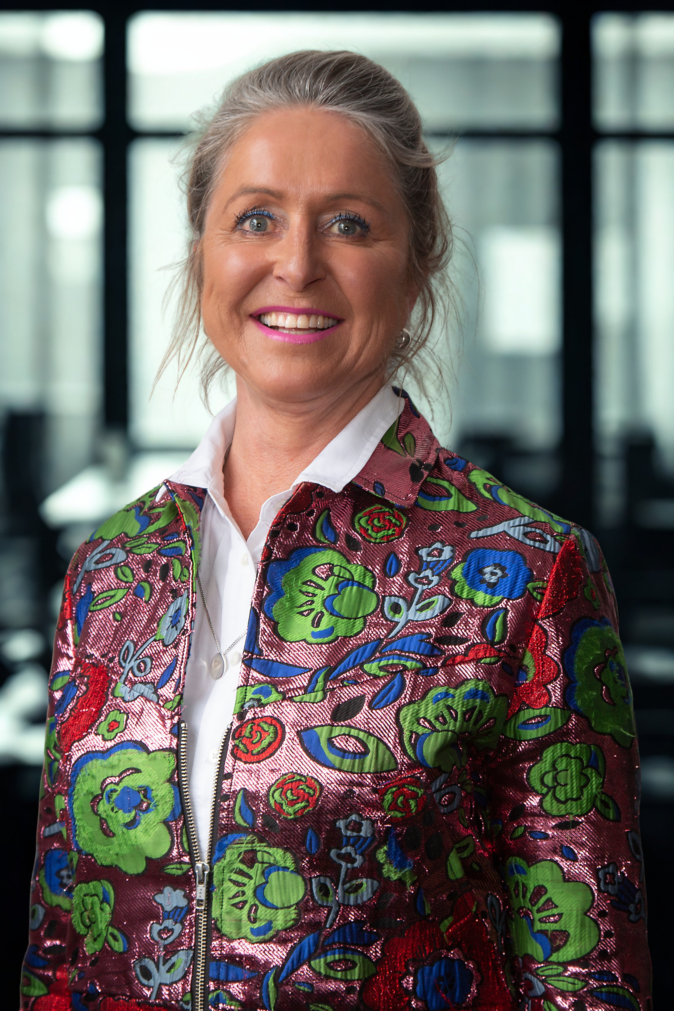 A smiling woman with gray hair tied back, wearing a colorful embroidered jacket and a white shirt, standing indoors with a blurred background of windows.