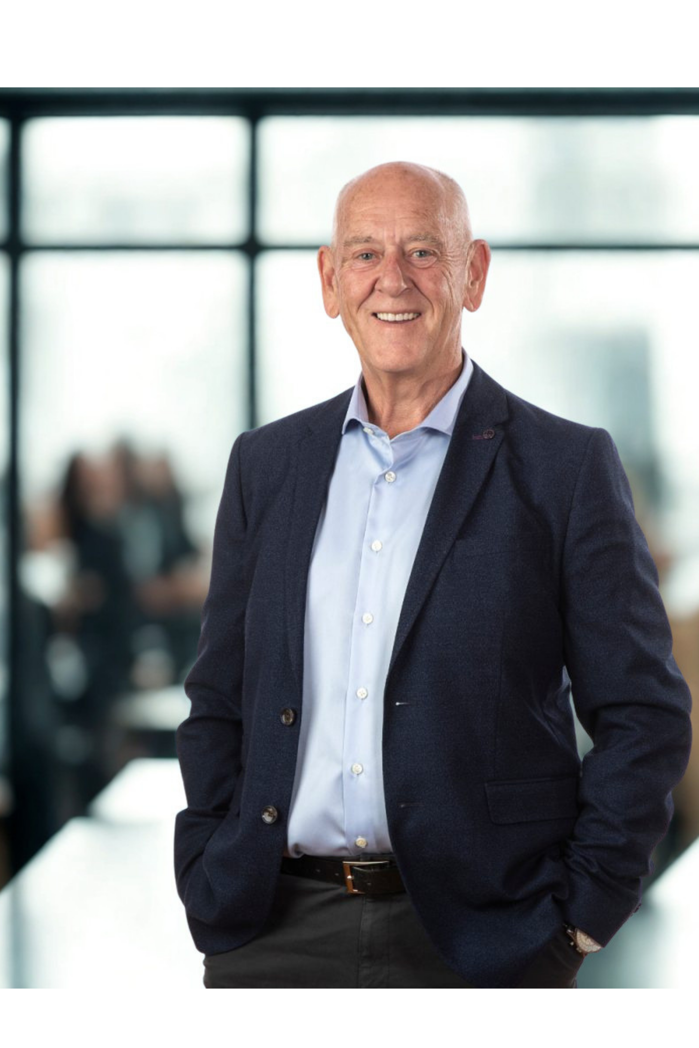 A smiling elderly man in a dark gray suit and light blue shirt standing in an office with large windows in the background.