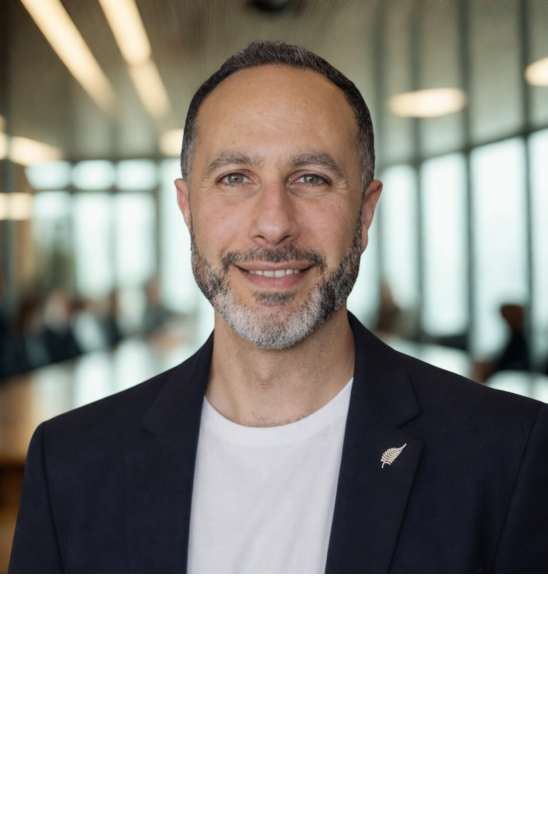 Close-up of a middle-aged man with short dark hair, grey beard, wearing a black blazer with a pin, smiling, in a professional setting with blurred background of a modern office or conference room.