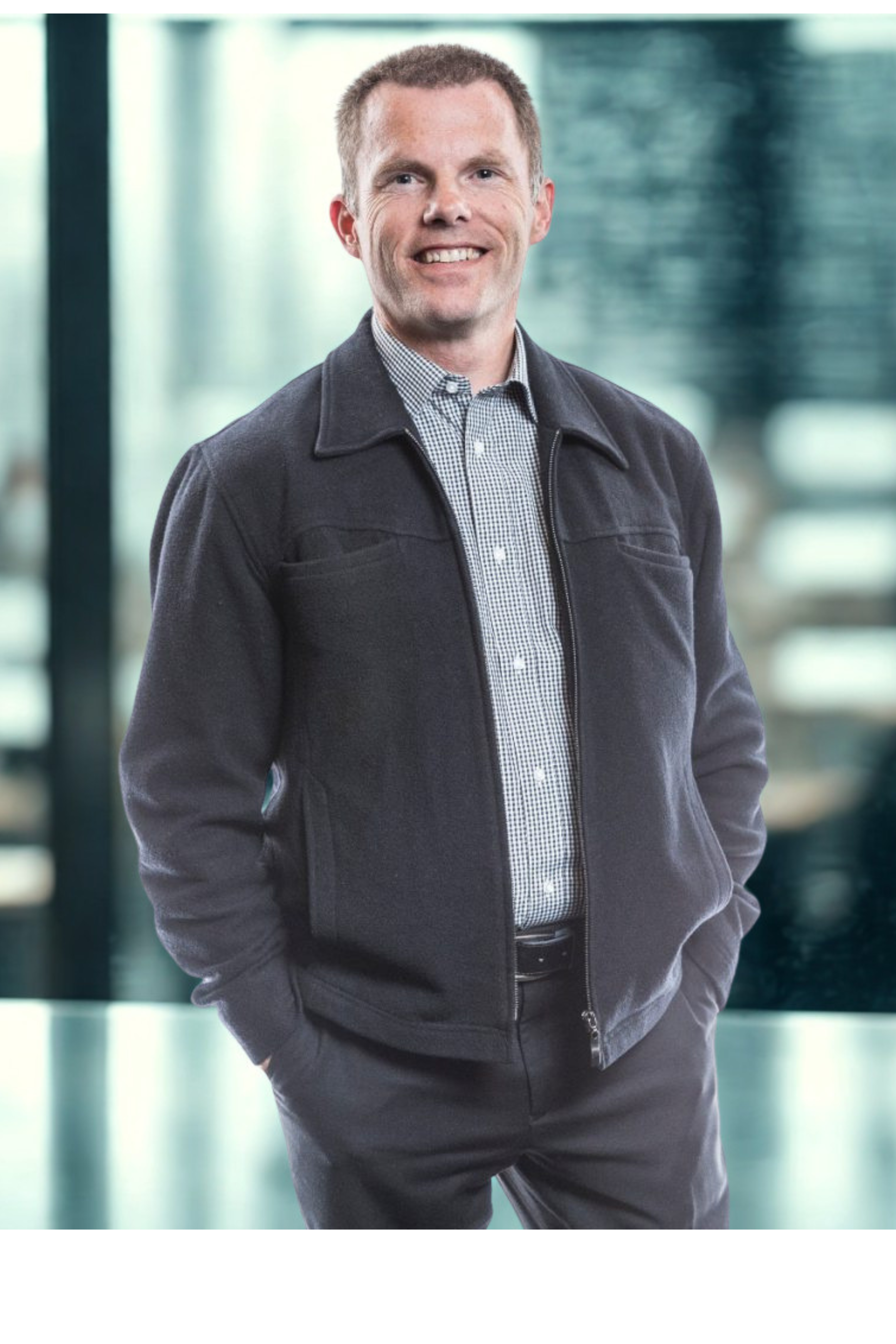 A man with short hair and a broad smile wearing a checked shirt under a dark zip-up jacket, standing with hands in pockets in a modern indoor setting with large windows and blurred background.