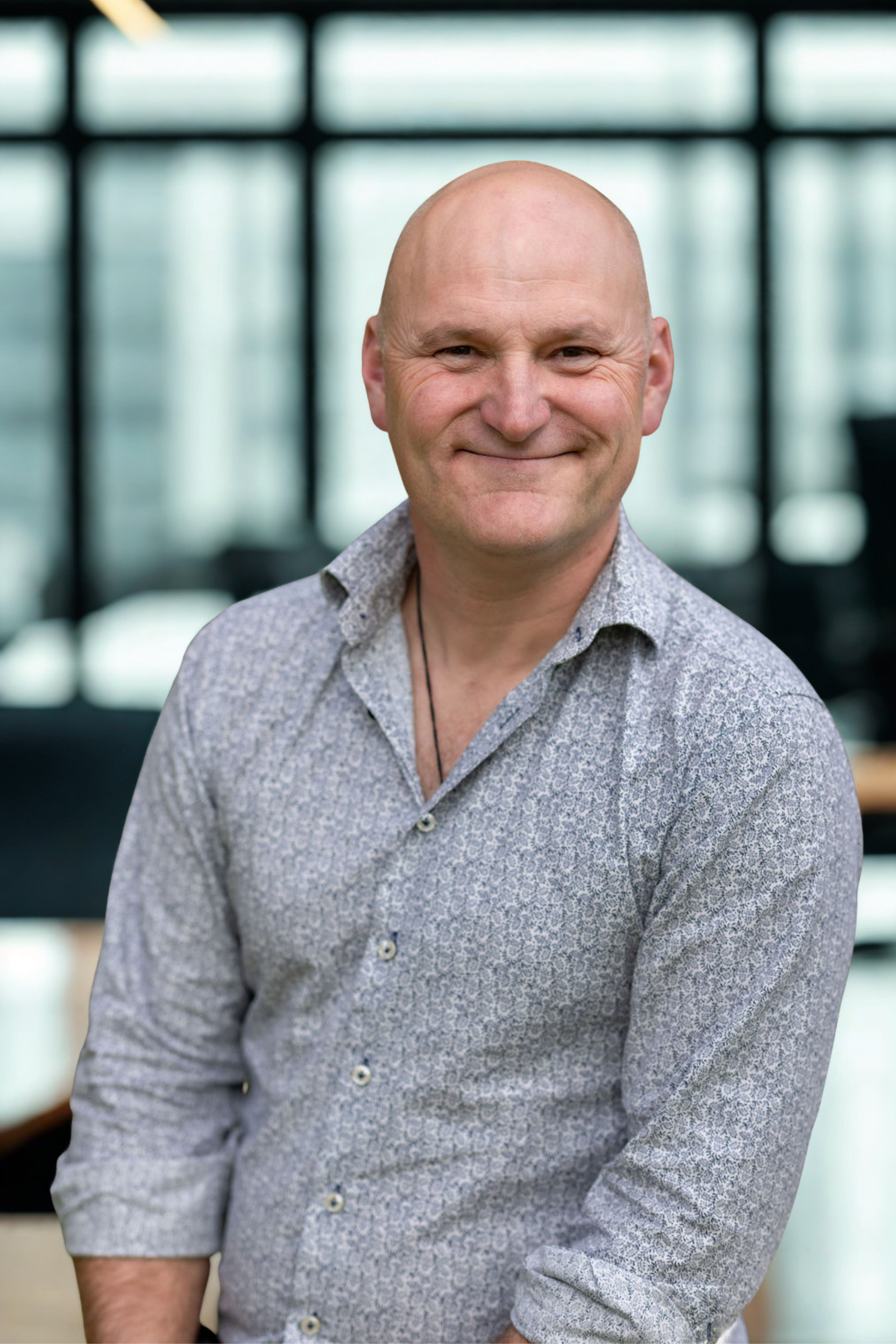 A middle-aged man with a bald head smiling, wearing a light patterned shirt, in a modern building with large glass windows in the background.