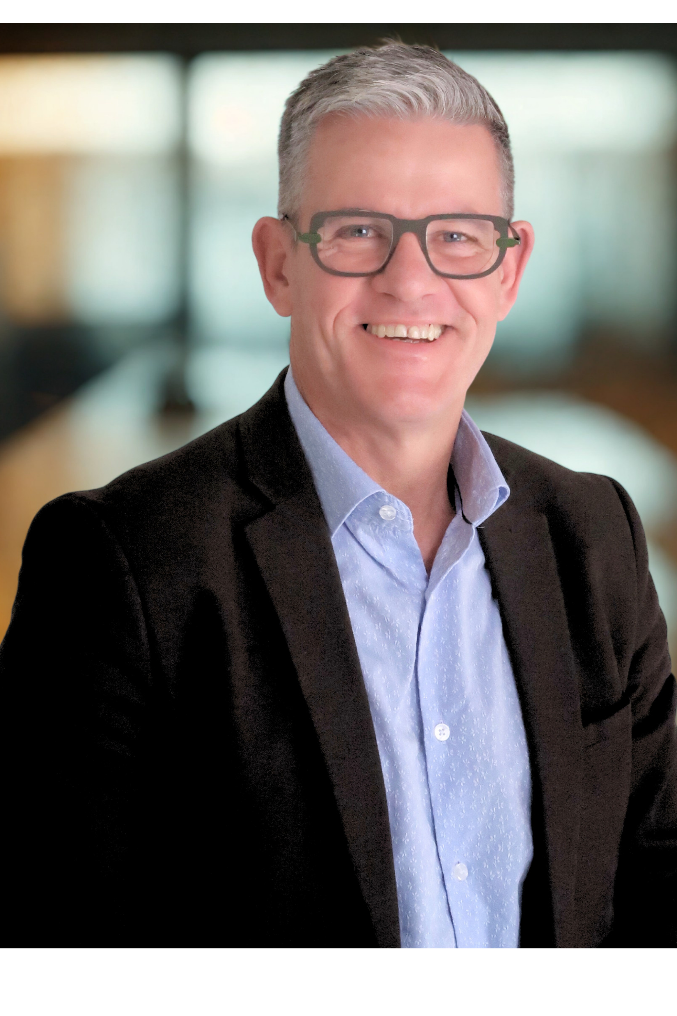 Smiling man with gray hair and glasses, wearing a black blazer and a light blue shirt, in a professional setting with blurred background.