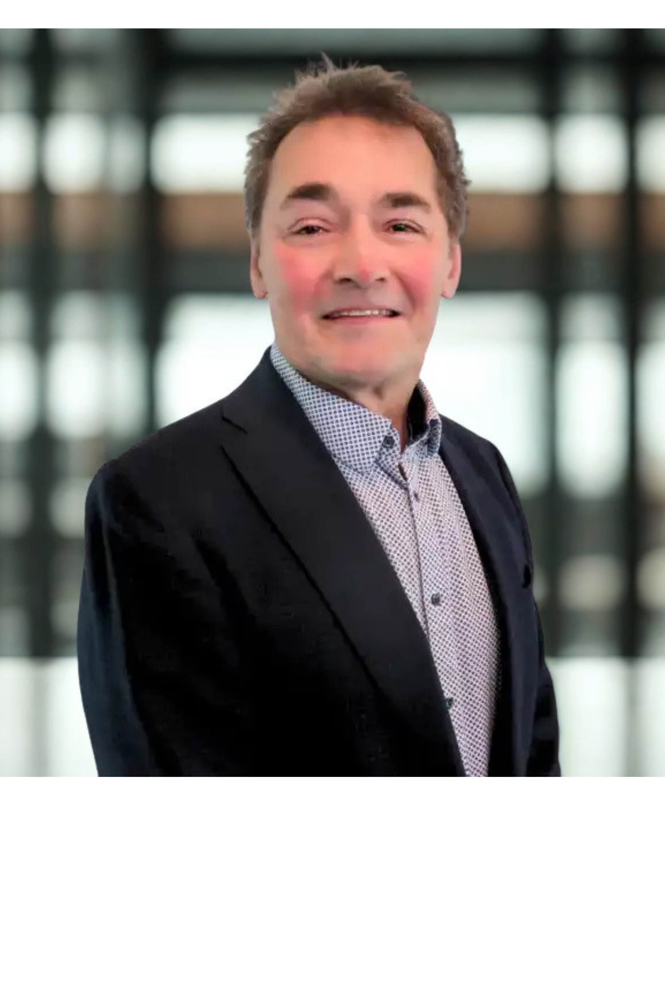 Close-up of a smiling man in a formal suit and shirt, standing in a modern building with large, blurred windows in the background.