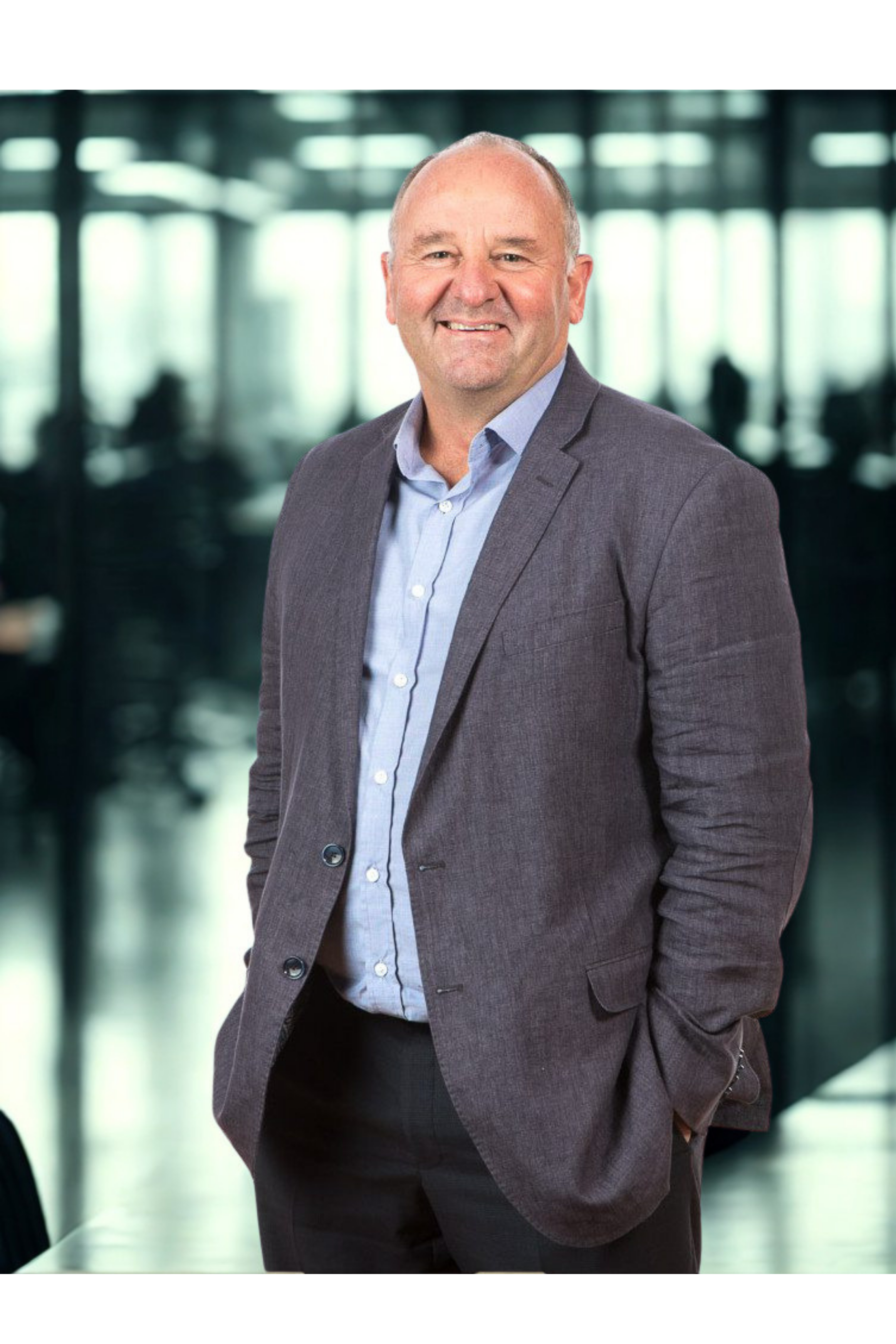A middle-aged man with a bald head, light skin, and wearing a dark gray blazer over a light blue dress shirt, standing in a brightly lit modern office lobby with glass walls and silhouettes of people in the background.
