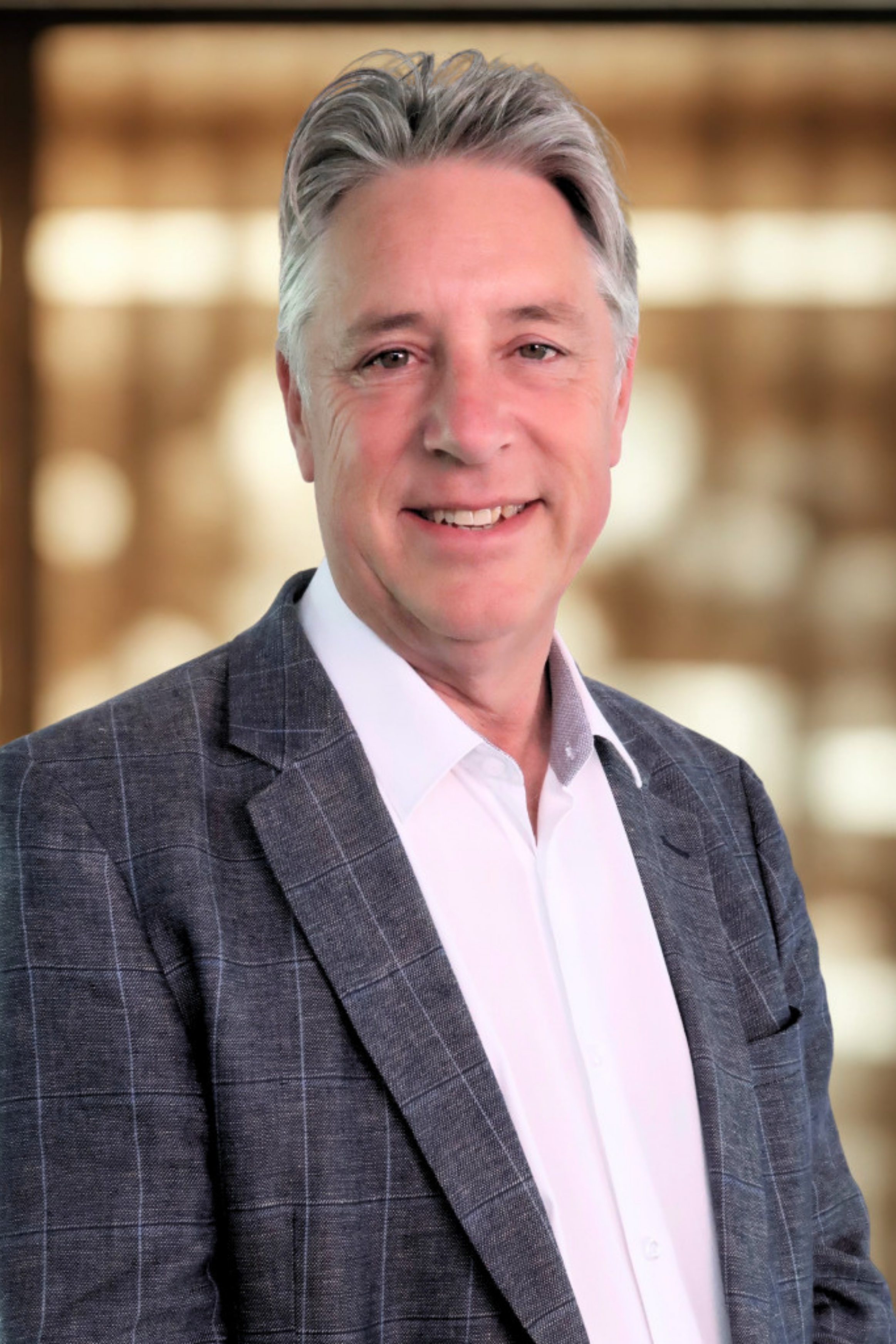A man with gray hair and a friendly smile wearing a checked gray suit jacket and a white shirt, standing in front of a blurred background with warm tones.