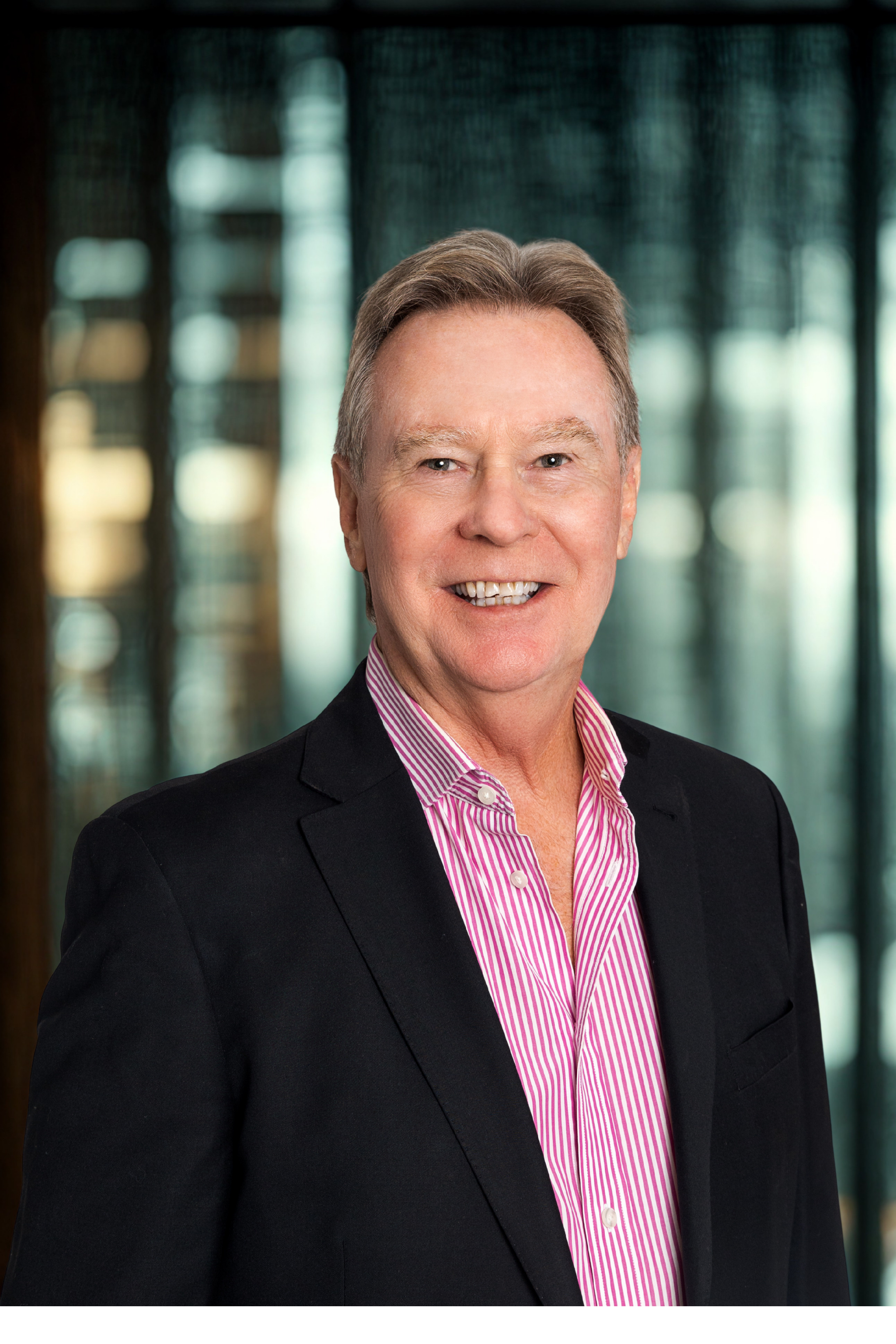 Head and shoulders portrait of a smiling middle-aged man with gray hair, wearing a black blazer and a pink and white striped button-down shirt, in front of a blurred indoor background with large windows.