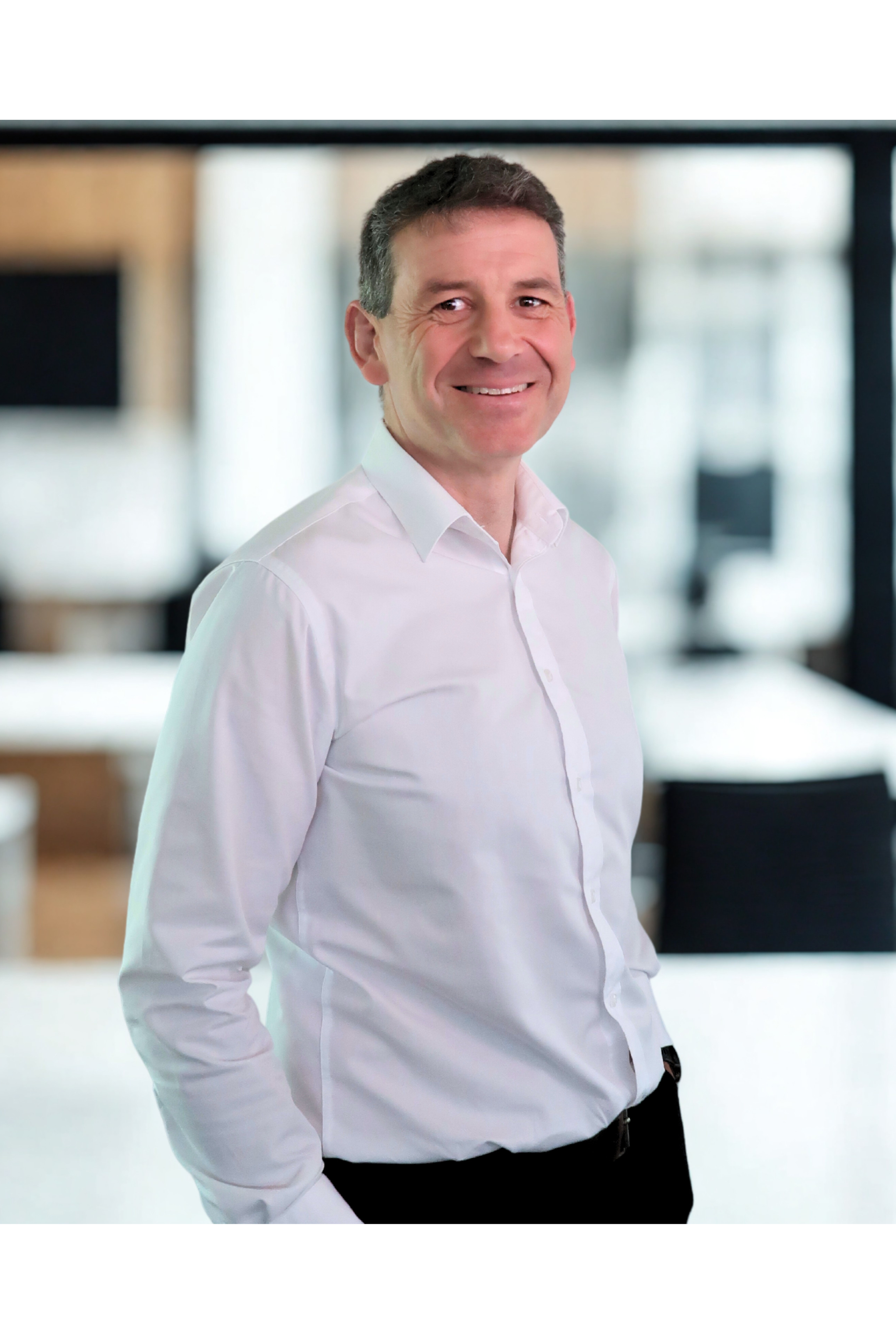 A smiling man in a white dress shirt standing indoors with a blurred background of windows and furniture.