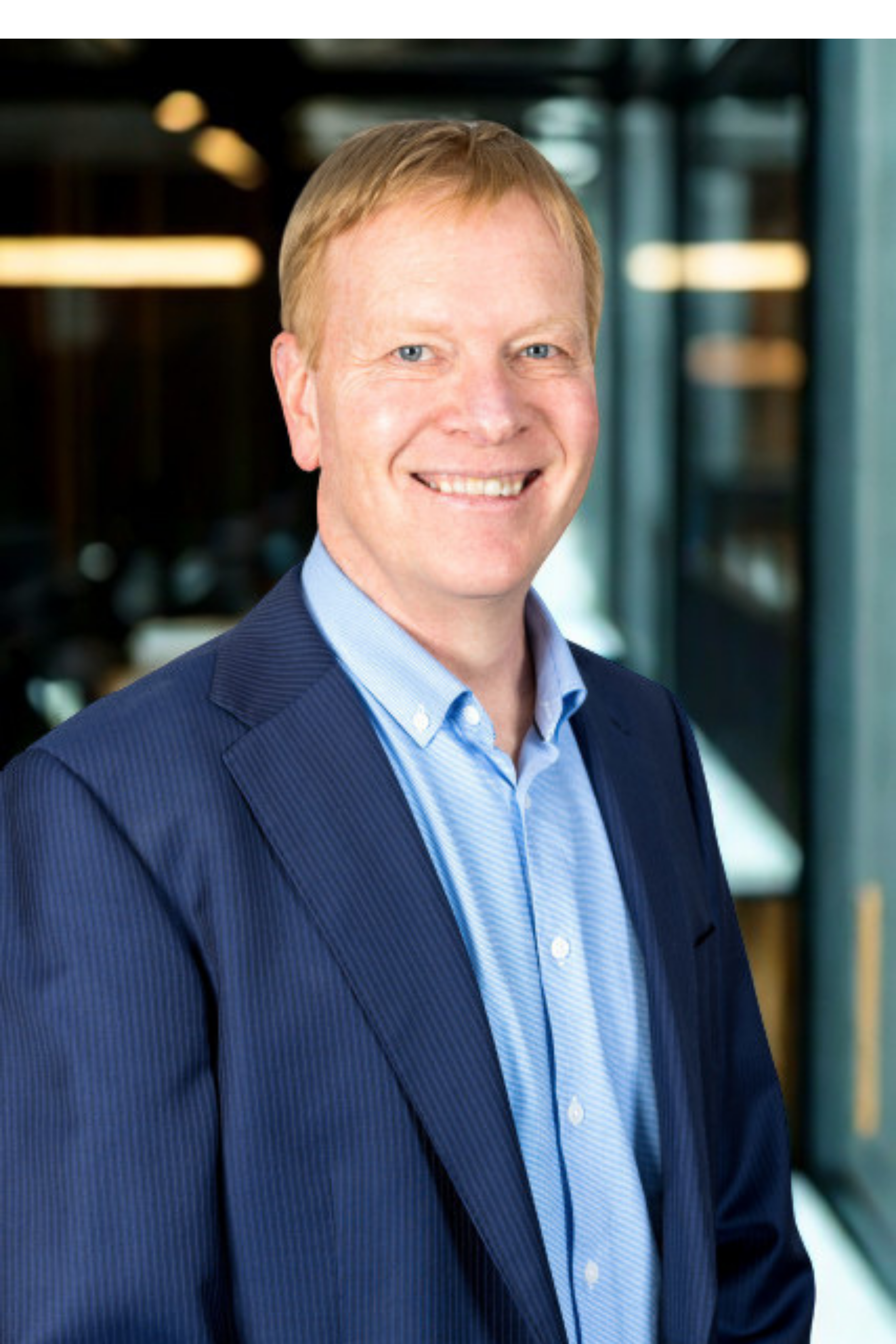 Portrait of a smiling man in a blue suit and light blue shirt indoors, with a blurred background of bookshelves and windows.