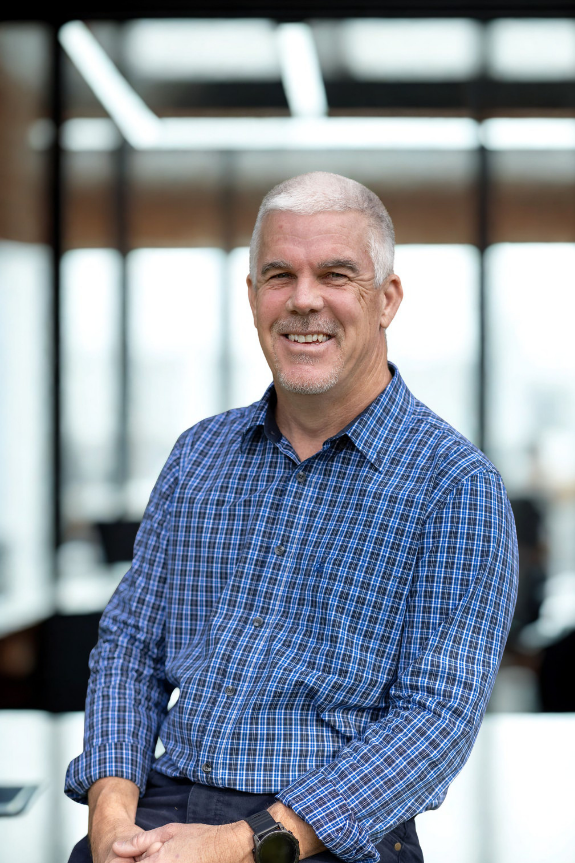A middle-aged man with short gray hair styled neatly, smiling, wearing a blue checkered shirt with sleeves rolled up, standing in a modern indoor space with large windows and a wooden ceiling.