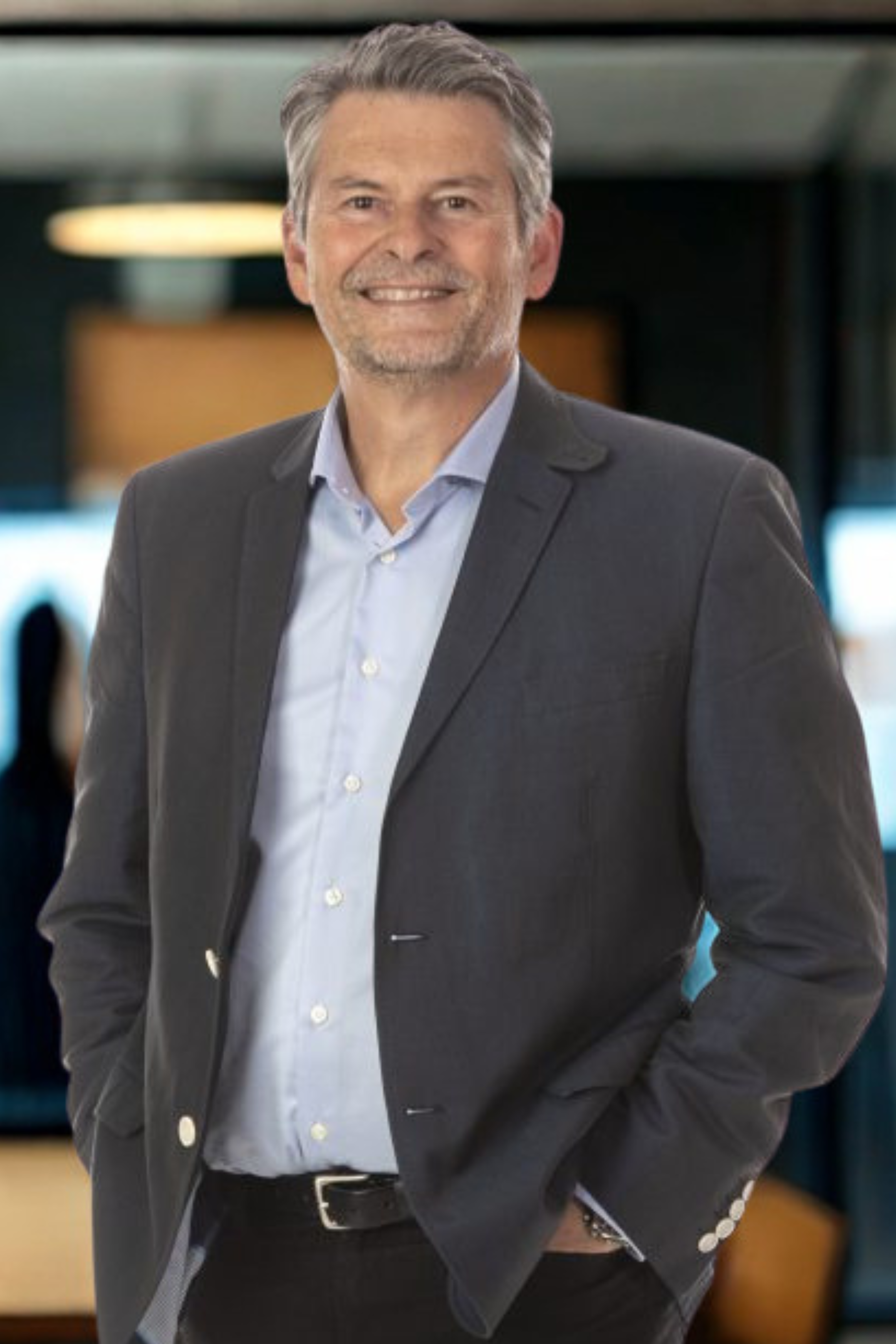 A middle-aged man with gray hair and a beard, dressed in a dark suit jacket and a light-colored shirt, smiling in an indoor setting.