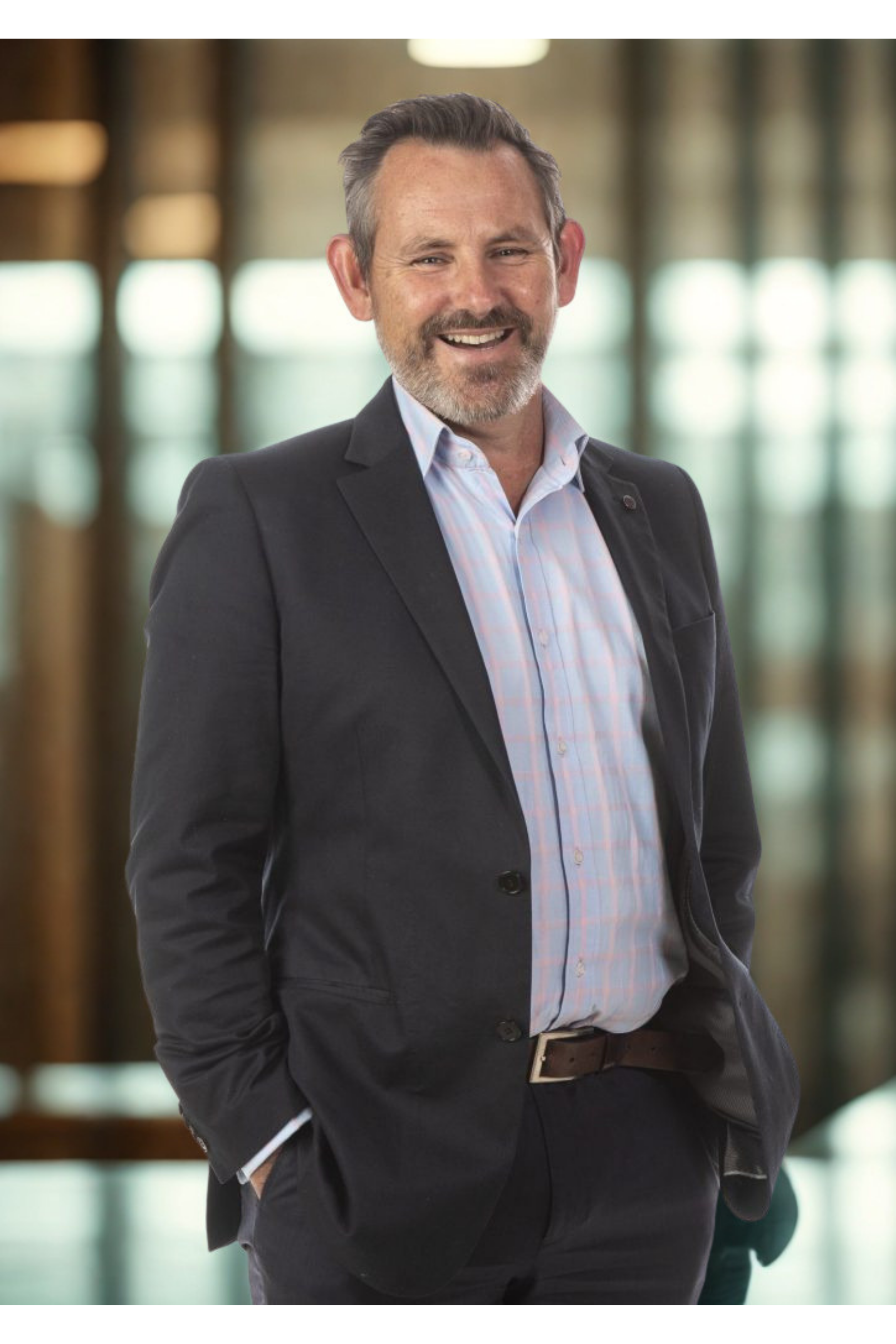A middle-aged man with gray hair and a beard smiling, dressed in a dark blazer and light shirt, standing indoors with blurred windows and natural light in the background.