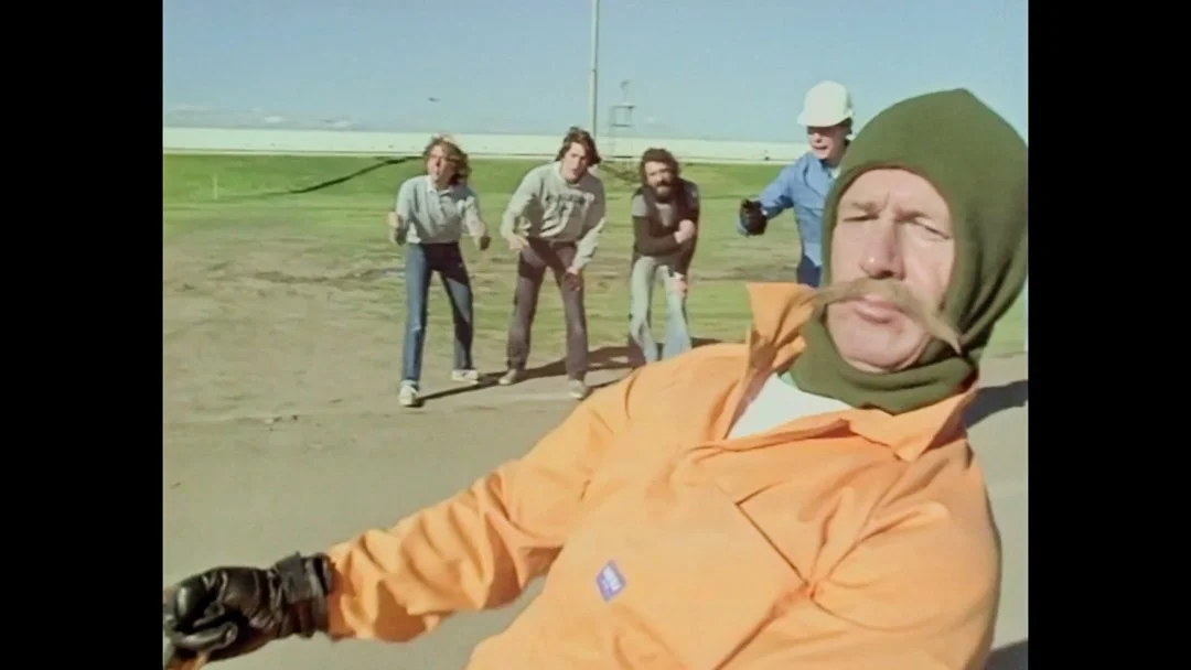 A man with a mustache and green headscarf taking a selfie on a deserted road with four people in the background, one wearing a construction helmet, during daytime.