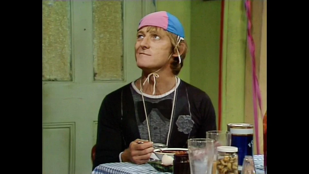 A man with blonde hair sitting at a table, wearing a colorful hat and a black shirt with a graphic print, inside a room with green walls and a door, with party decorations on the right side.