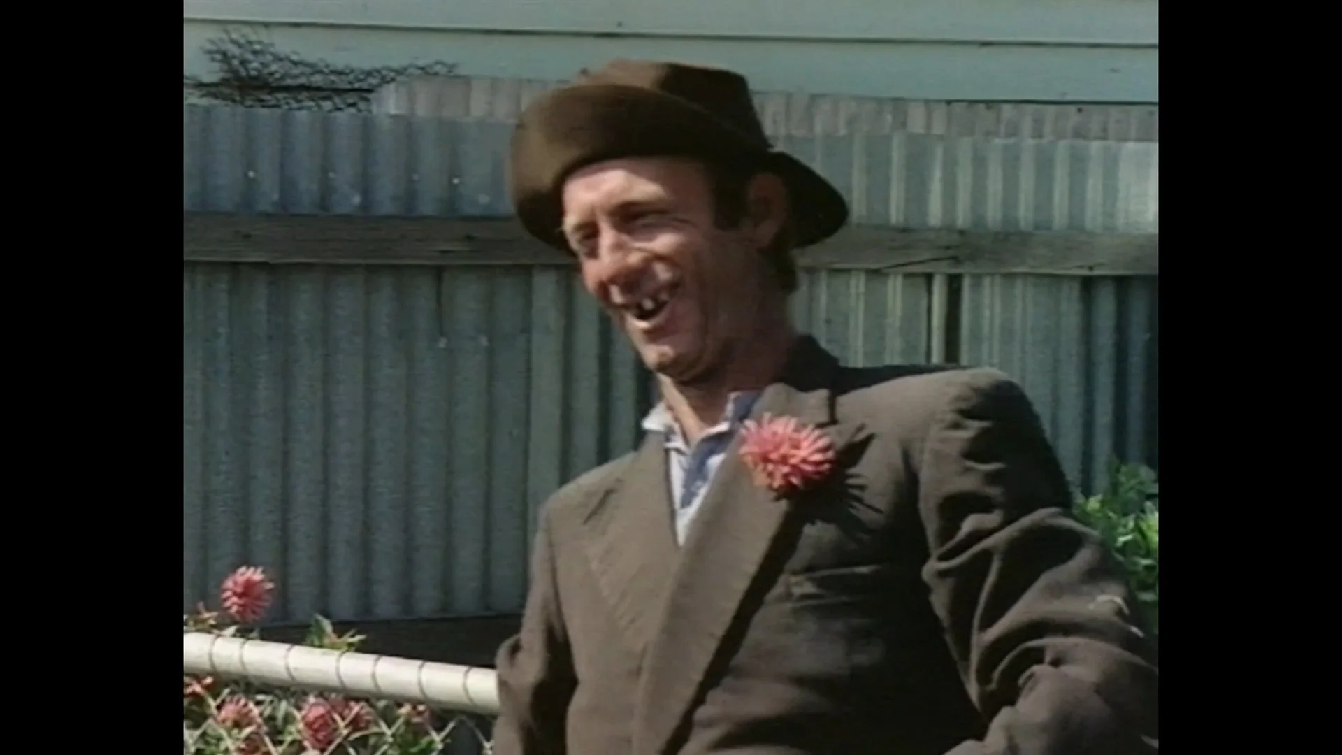 A man with a big smile, missing teeth, and a hat, dressed in a suit with a flower pinned to his lapel, standing outdoors near a fence.