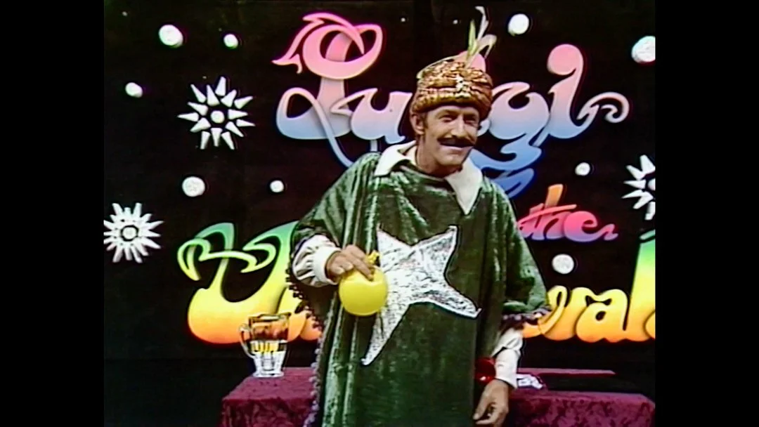 Man dressed as a chef with a turban, holding a yellow teapot, standing in front of a colorful sign that reads 'Chiquirri Badi Dios'.