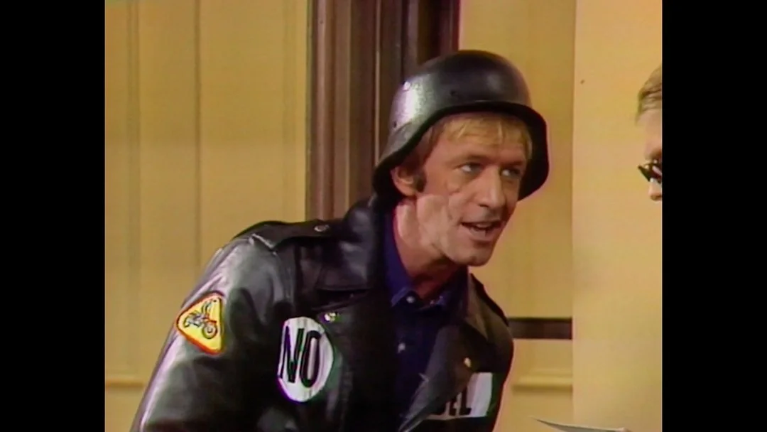 Nigel Lovelace is dressed in a helmet, leather jacket with patches, talking to someone in an indoor setting. 
