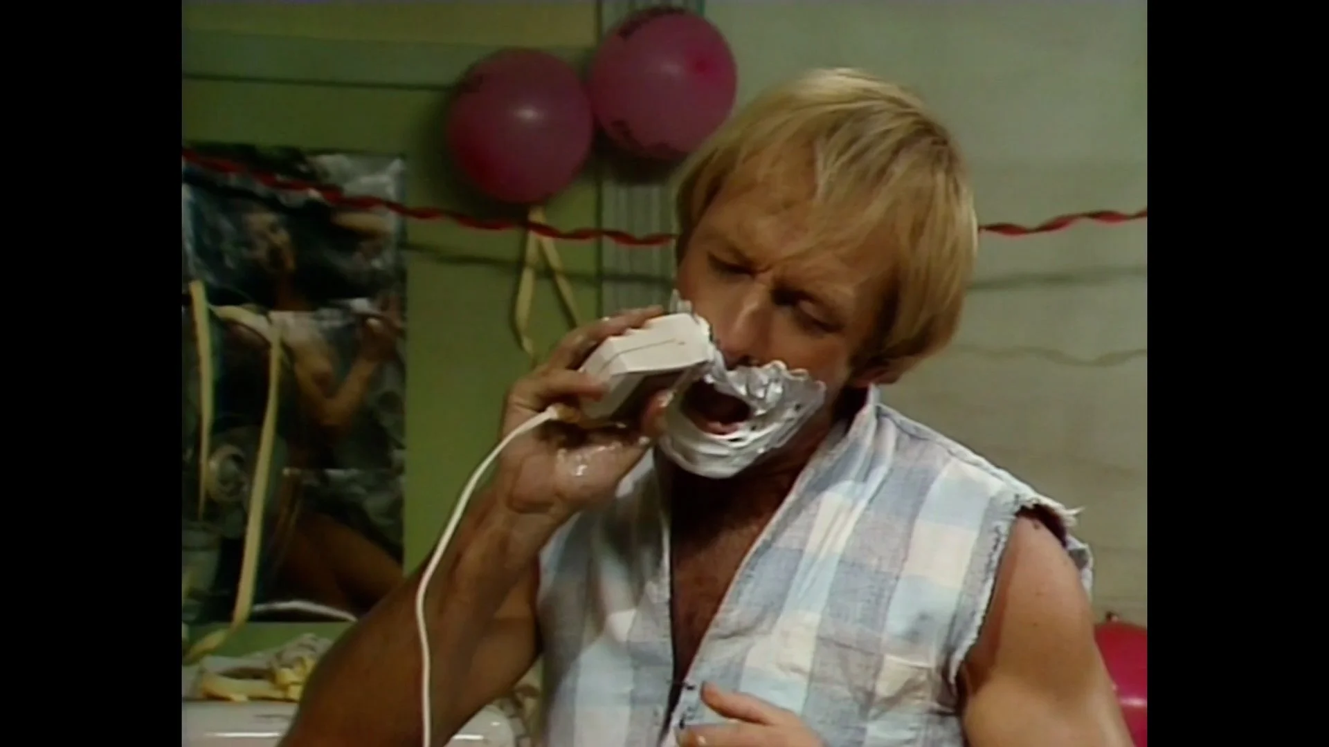 Hoges with blond hair, wearing a sleeveless plaid shirt, shaving with a shaver and foam