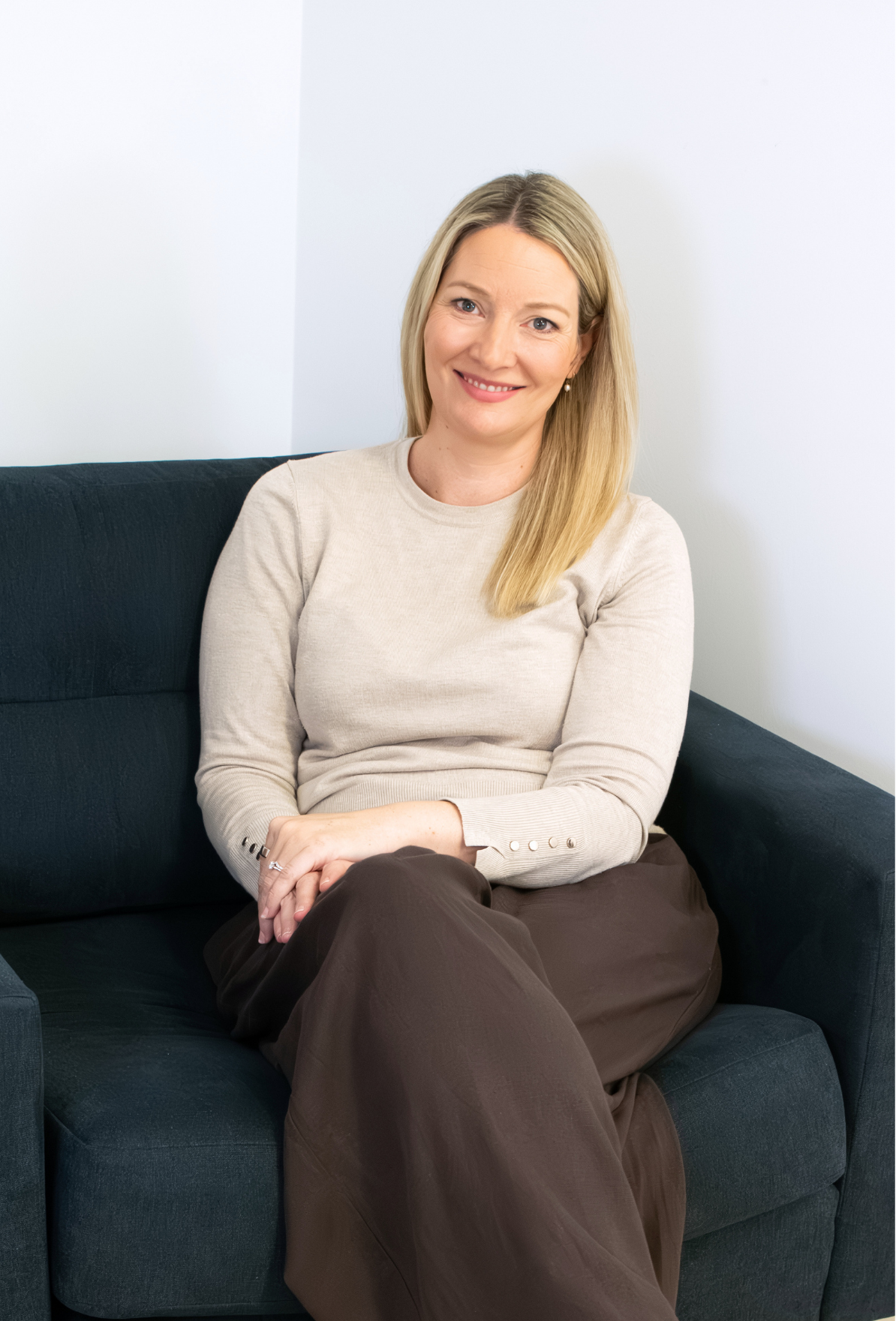 Smiling woman with blonde hair wearing a light coloured long sleeve top sitting in an armchair