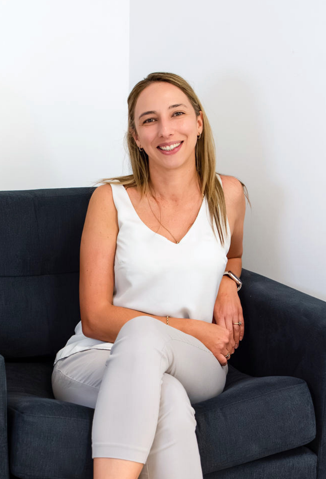 Smiling blonde woman wearing a sleeveless white blouse sitting in an armchair