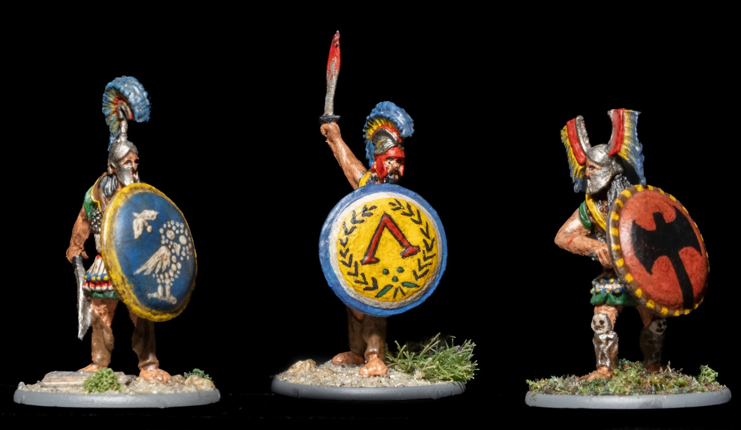 Miniature painted figurines of ancient warriors wearing colorful helmets and armor, holding shields and weapons, set against a black background.