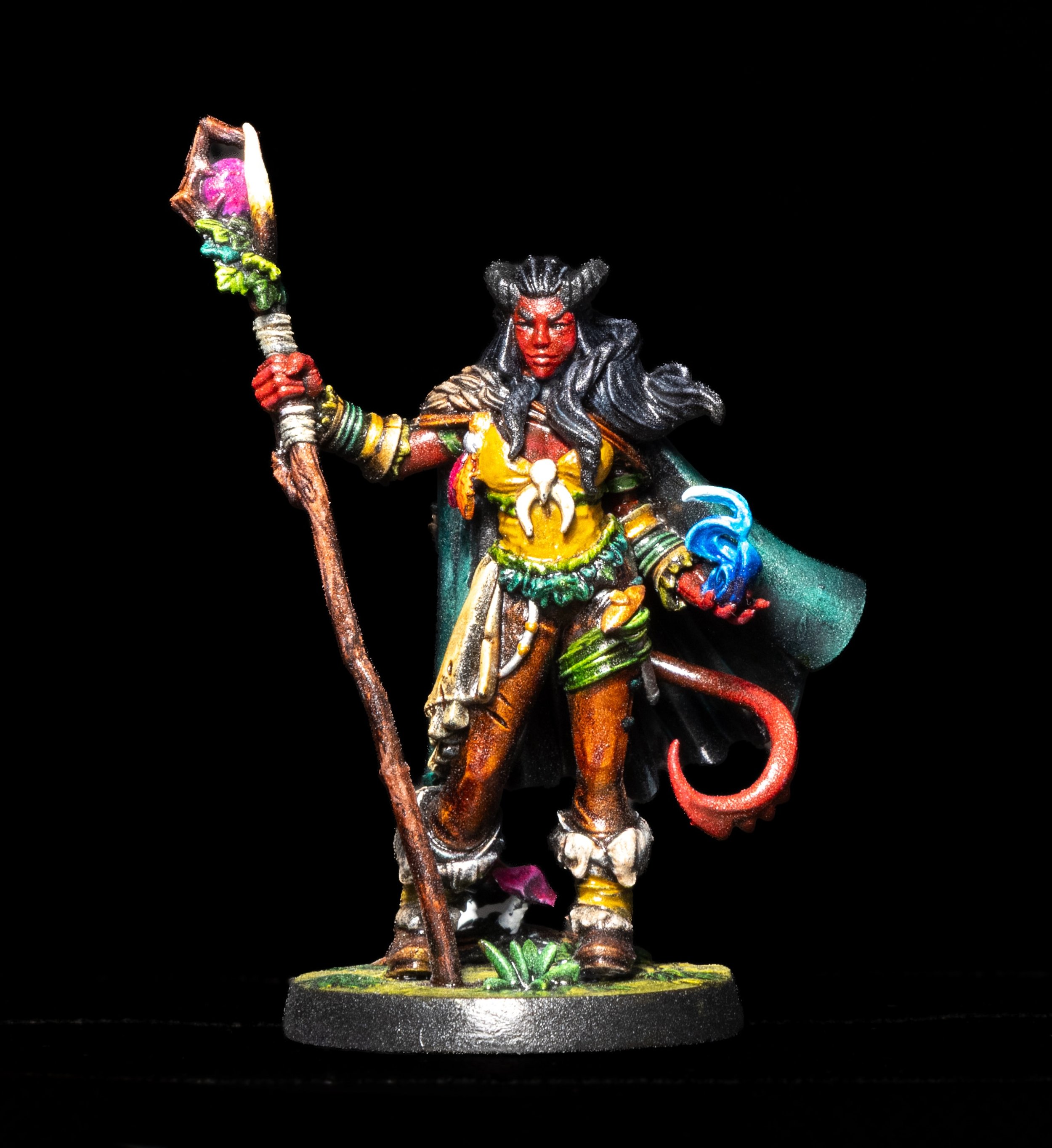 Colorful miniature figurine of a female warrior with dark hair, red face paint, and tribal clothing, holding a staff, standing on a round black base with green foliage accents.