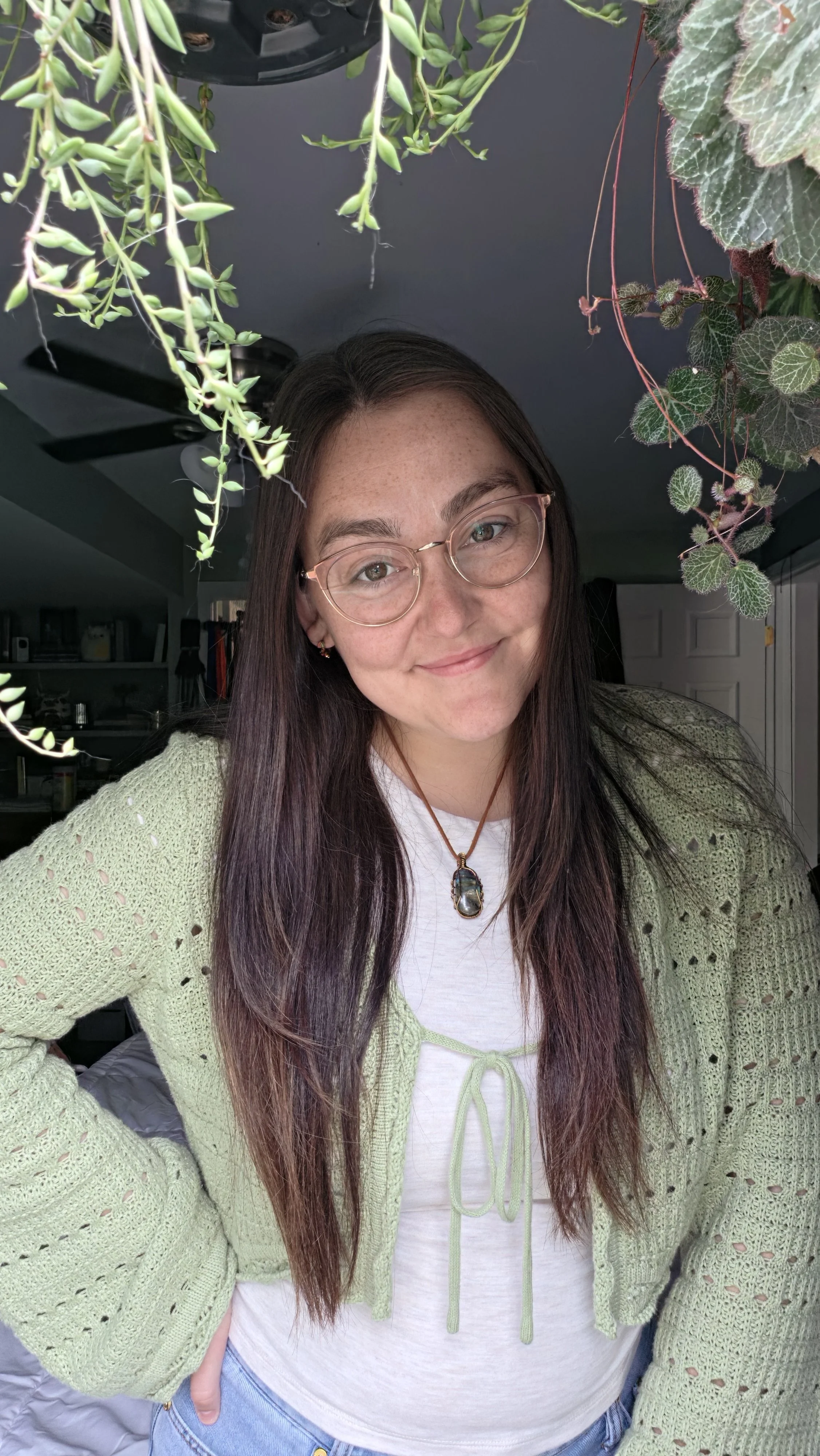 A young woman with long dark hair and glasses, smiling, wearing a light green knitted cardigan, a white T-shirt, jeans, and a necklace with a large pendant, standing indoors with hanging plants above her.