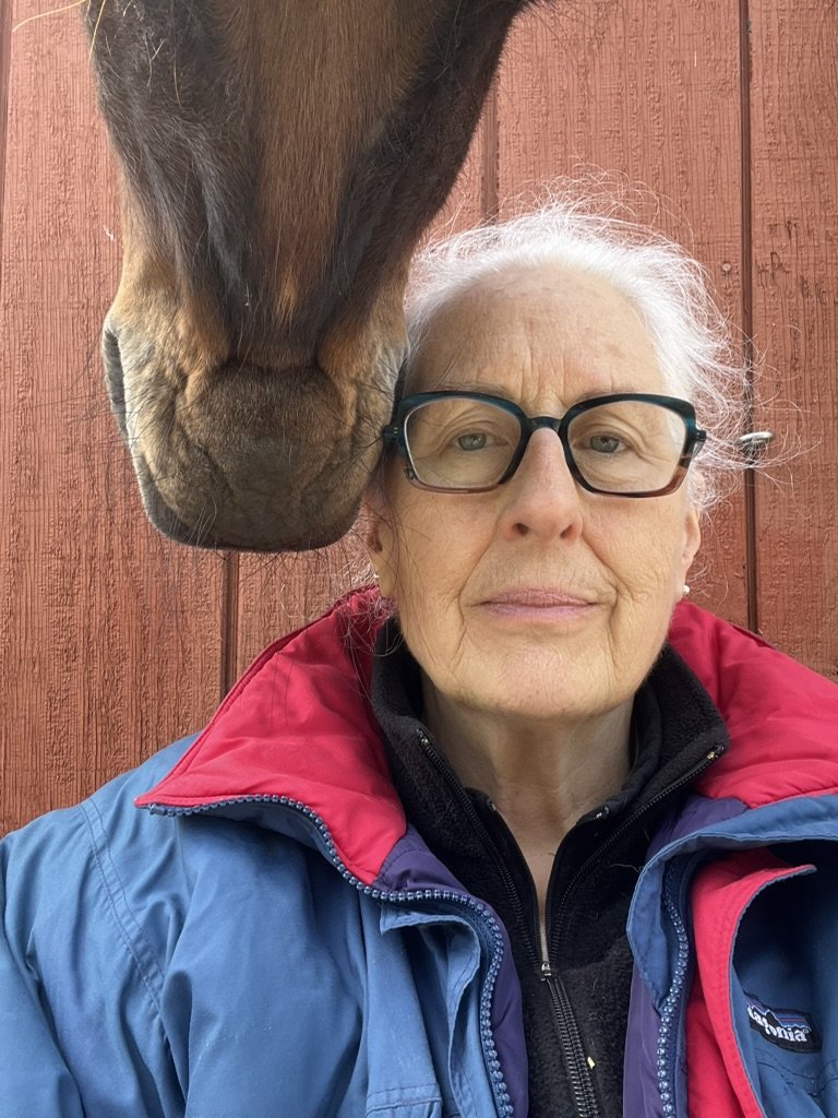 An elderly woman with white hair, glasses, and wearing a blue and red jacket, takes a selfie with a horse nearby, against a red wooden background.