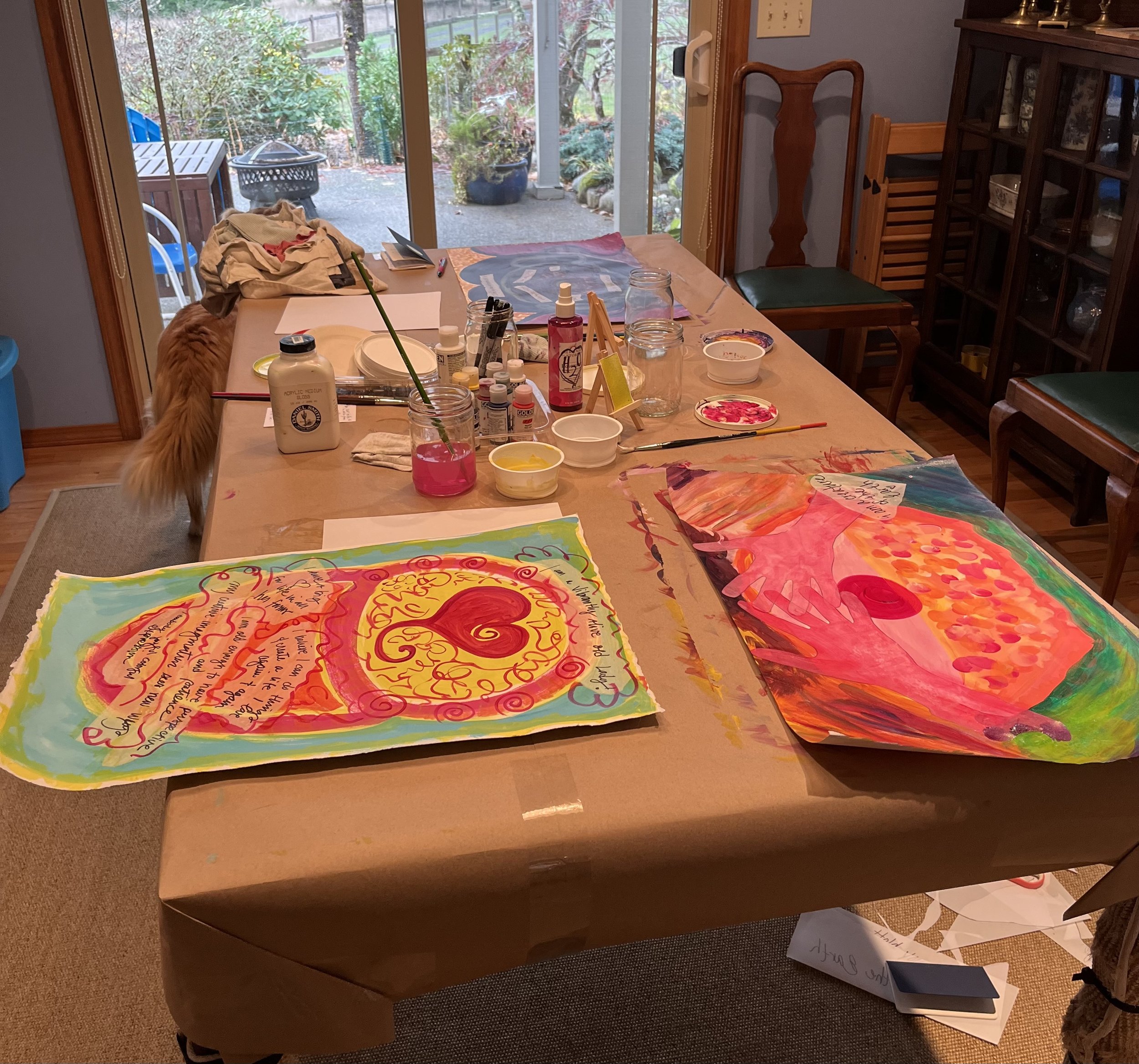 A cluttered large table with many colorful paintings and art supplies, including bottles of paint, brushes, and markers. The background shows a sliding glass door leading to a backyard with plants and trees.