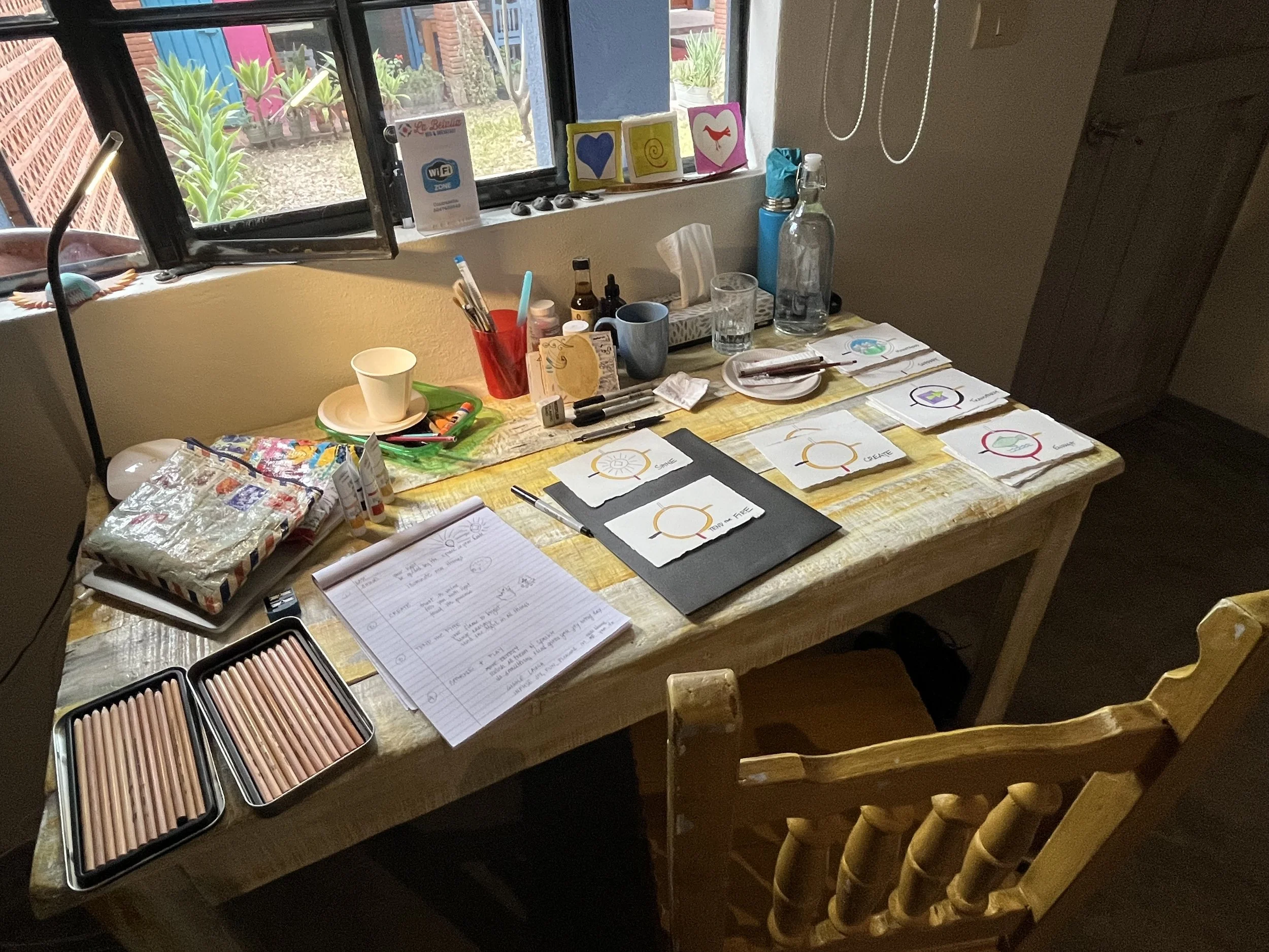 A cluttered wooden table with art supplies including colored pencils, markers, notebooks, and drawings. There are also some paper cups, a glass of water, and decorative items on the window sill. Outside the window, plants and a brick wall are visible.