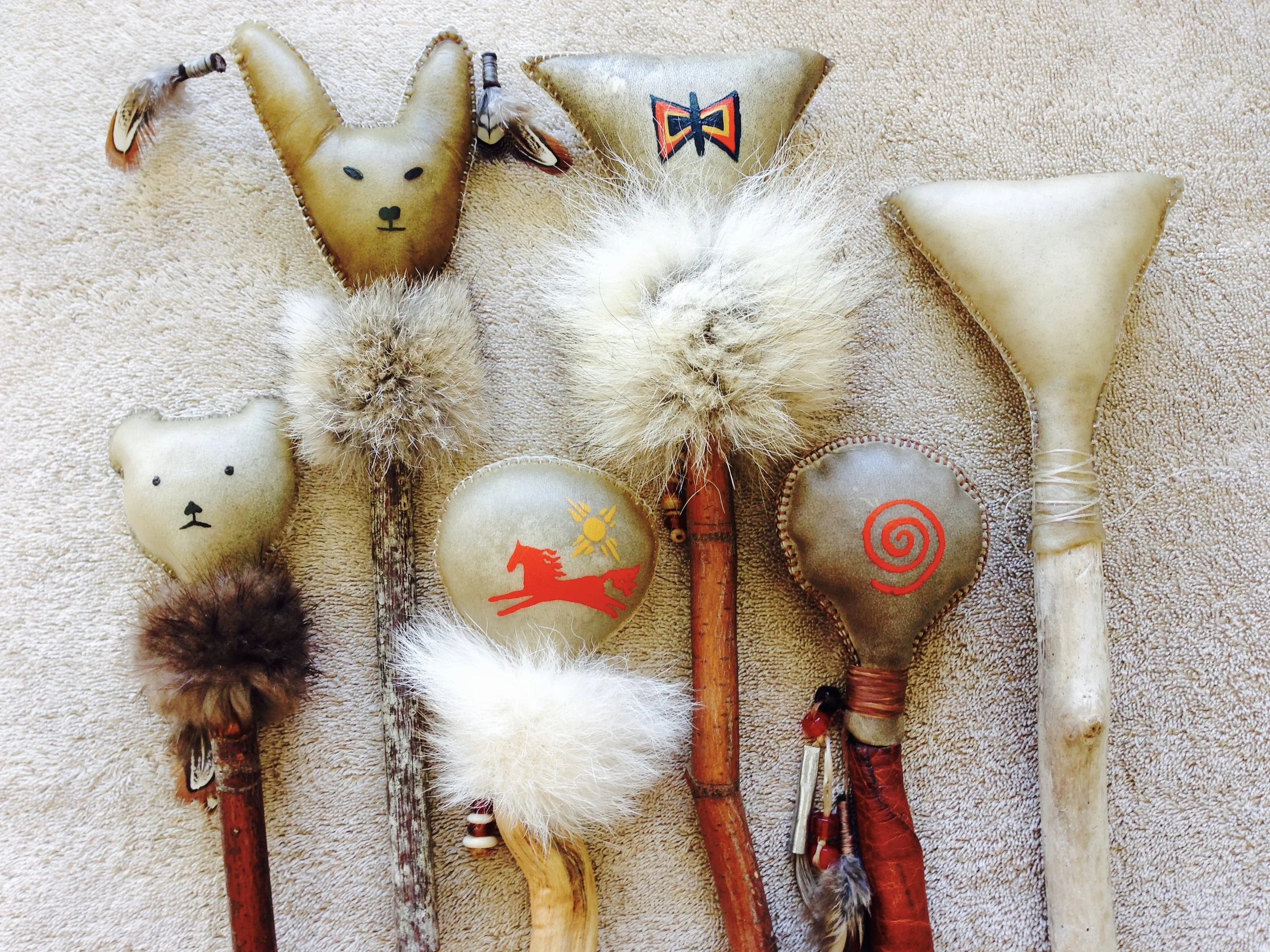 A collection of decorative spoons with animal face designs, feathers, and symbols, laid out on a beige textured surface.