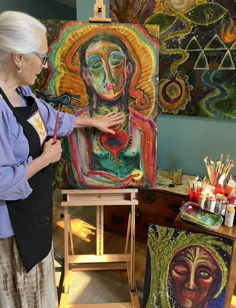 An artist with gray hair, glasses, and a black apron is painting a colorful portrait of a woman on a canvas. The artist is holding a paintbrush and touching the painted woman's chest. There are additional abstract and colorful artworks in the background and art supplies on a table nearby.
