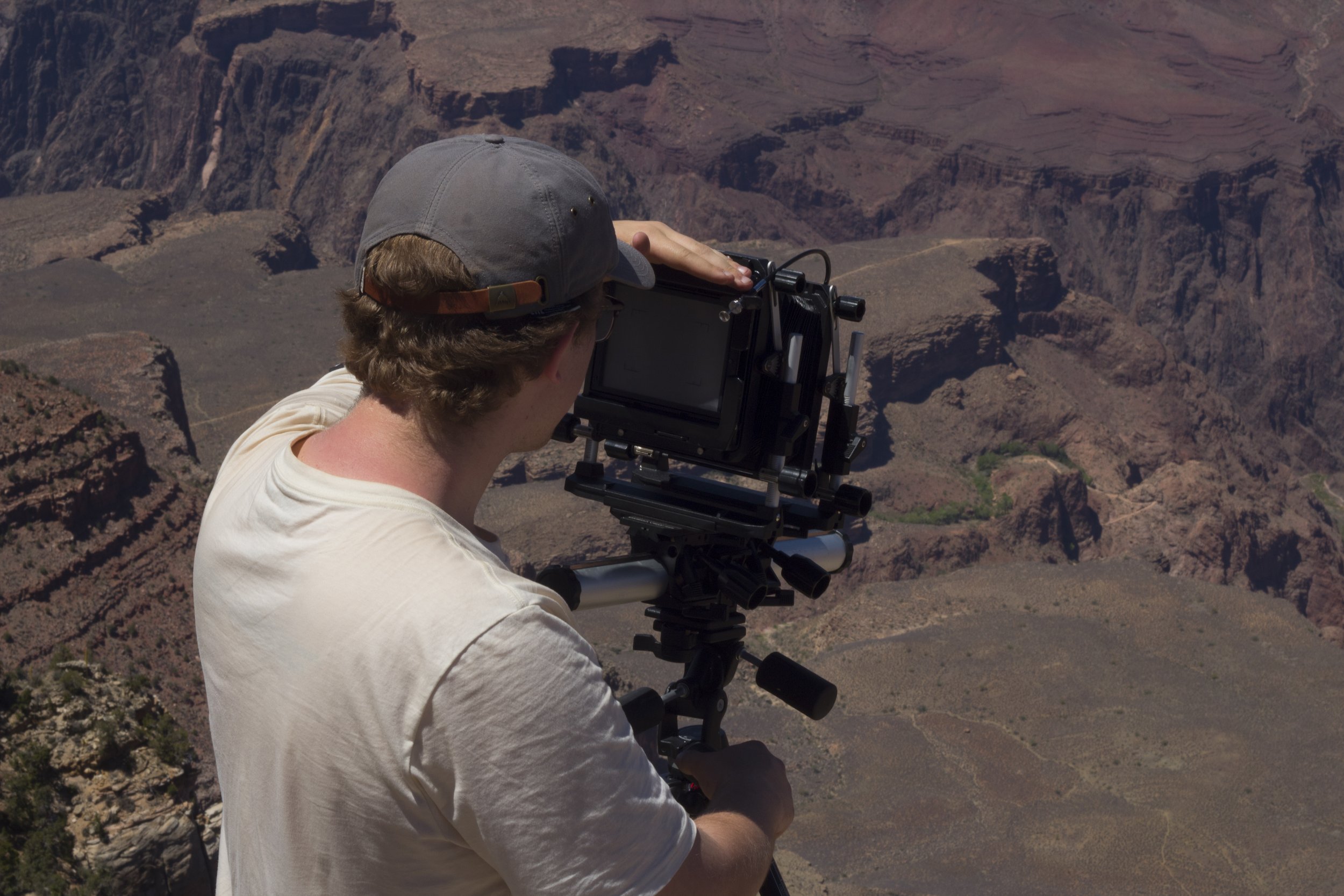 A person wearing a gray baseball cap and white shirt operating a professional camera mounted on a tripod, capturing a scenic view of the Grand Canyon with its layered rock formations and deep valleys.