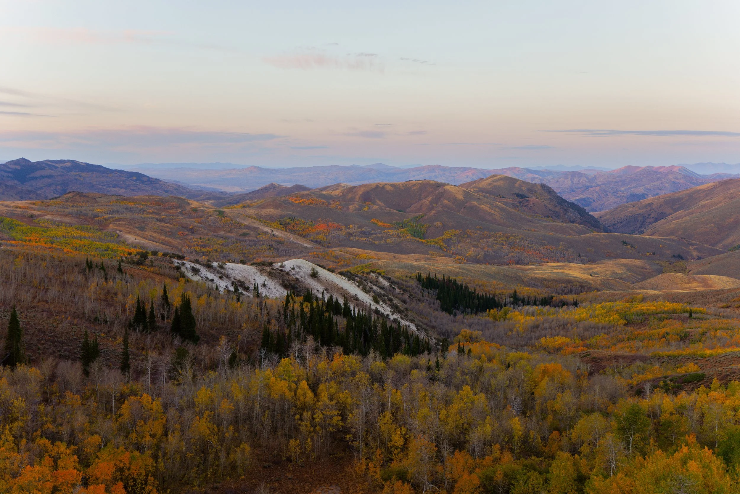 A vast landscape of rolling hills and mountains during fall, covered in colorful autumn foliage, under a partly cloudy sky.