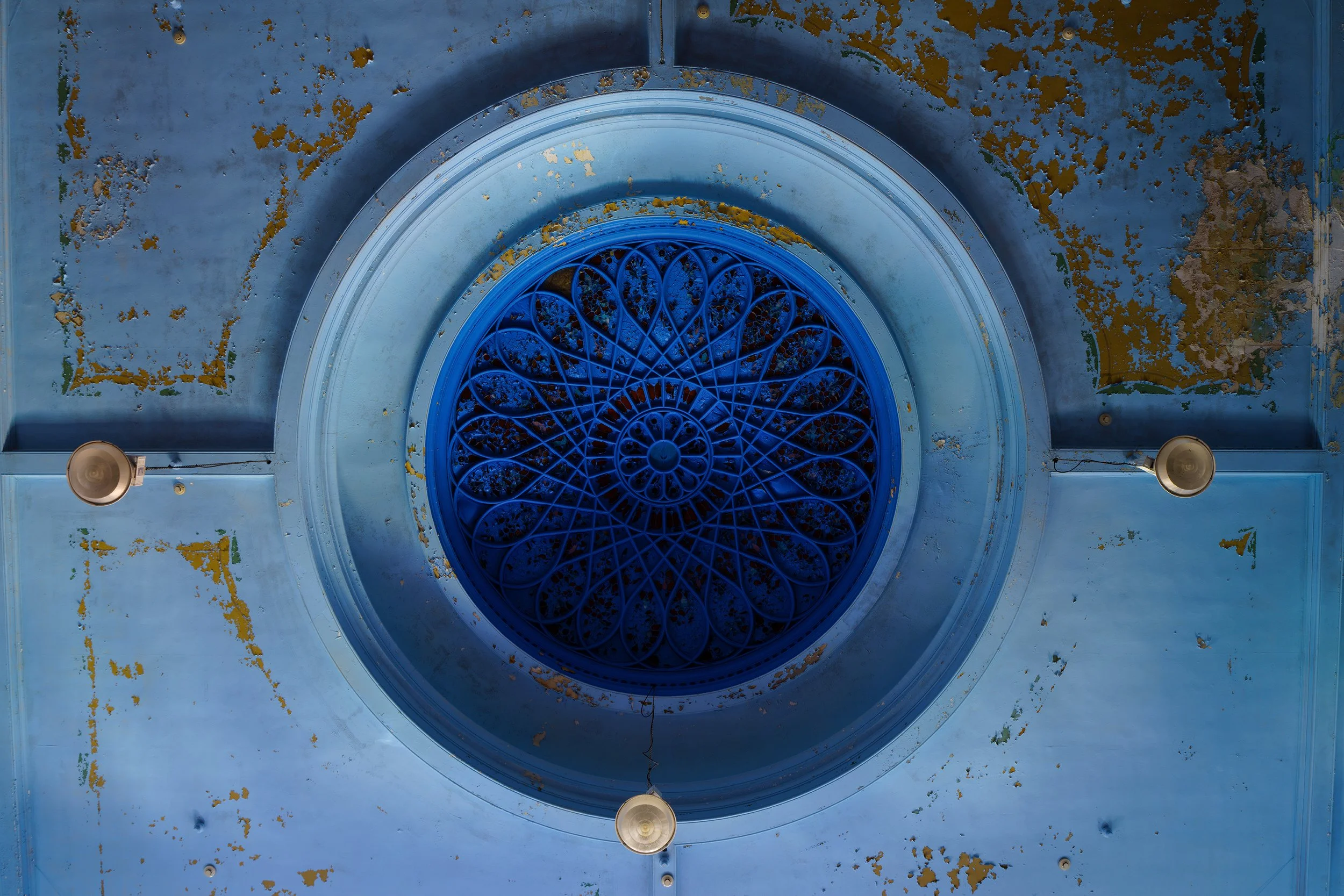 A ceiling with a large circular blue stained glass window featuring intricate geometric designs. The ceiling shows signs of age with peeling paint and worn patches. There are three beige or cream-colored light fixtures hanging from the ceiling.