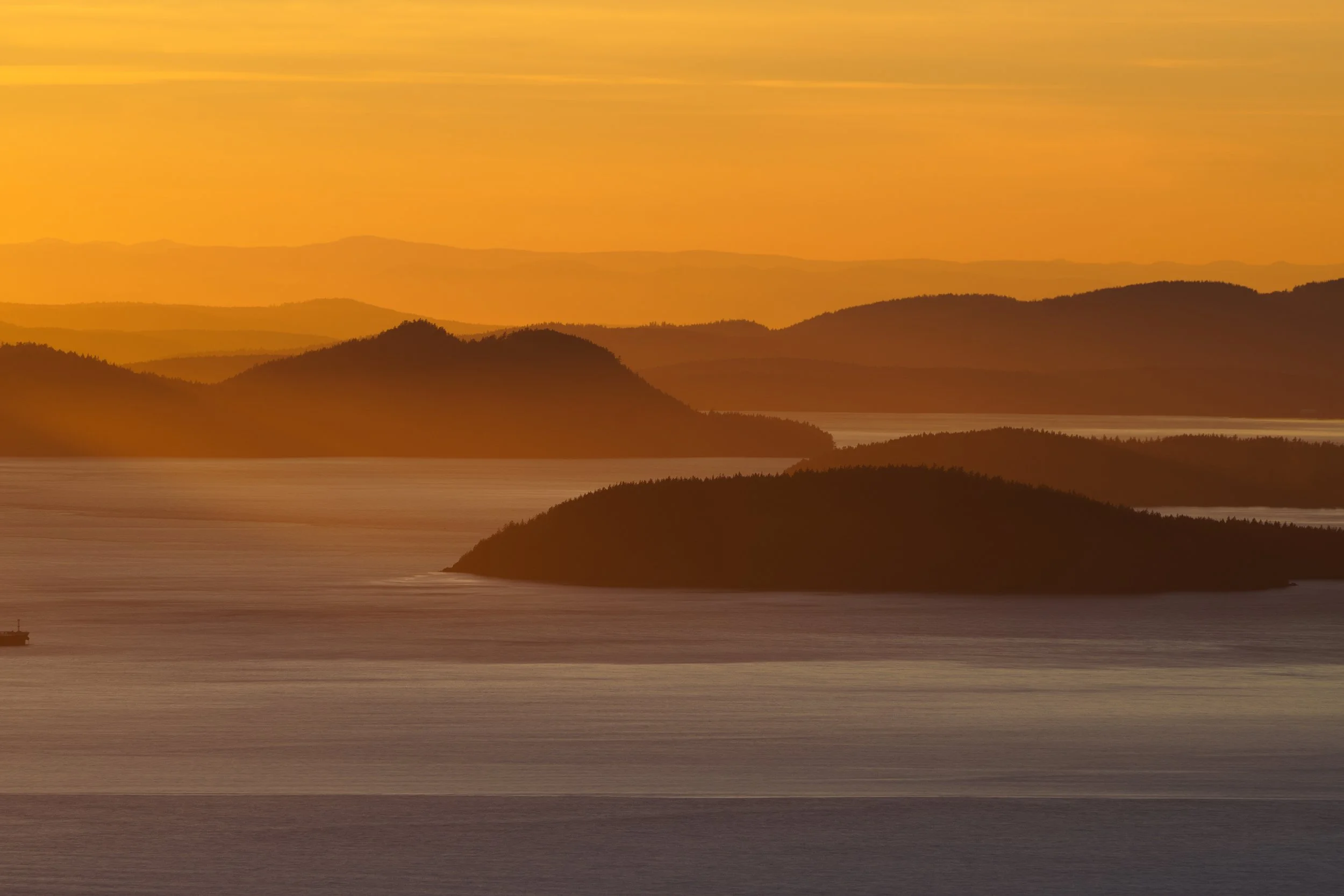 Sunset over a body of water with silhouetted hills and mountains in the distance.