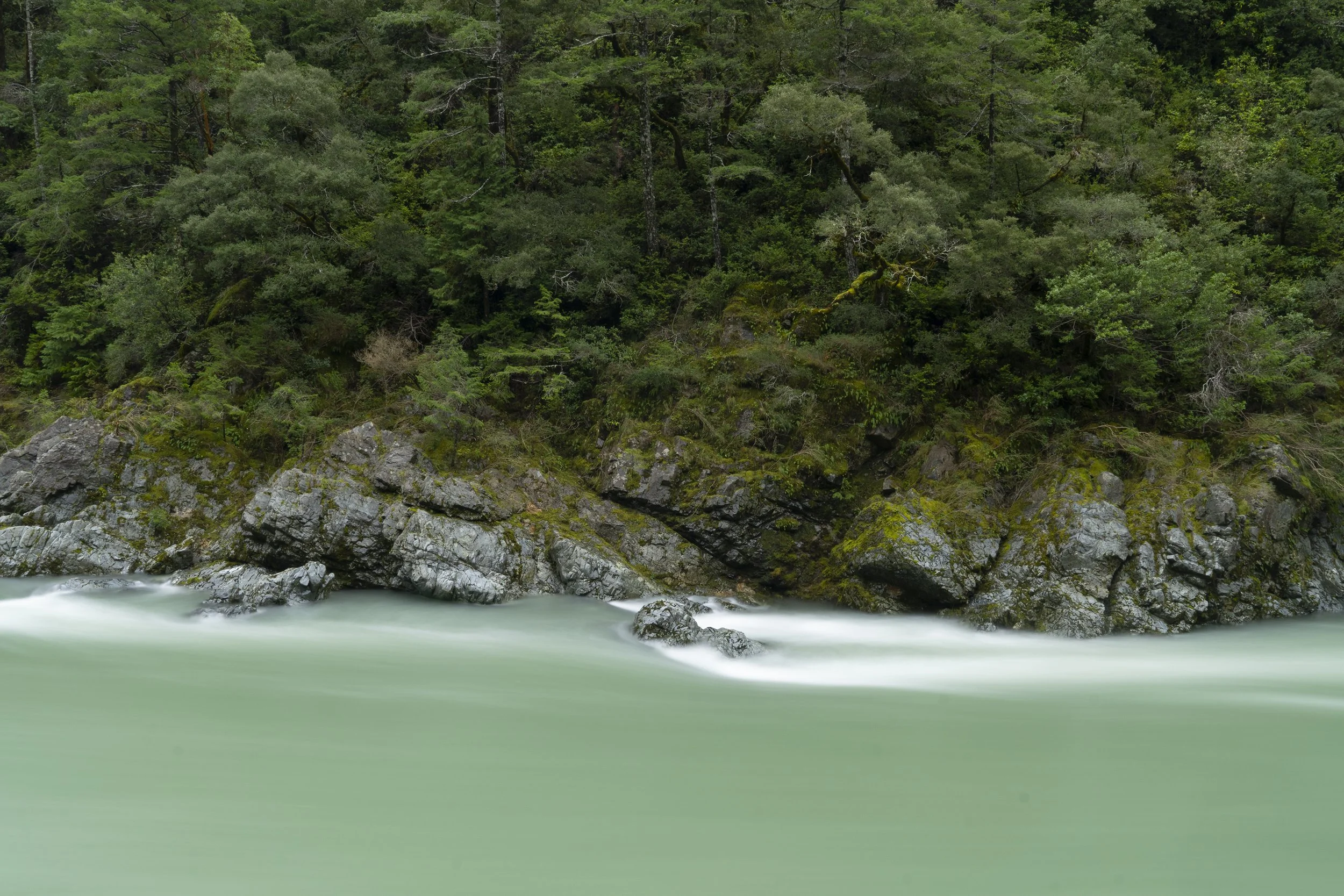 A river flowing through a lush green forest with trees on a hillside and rocks along the riverbank.
