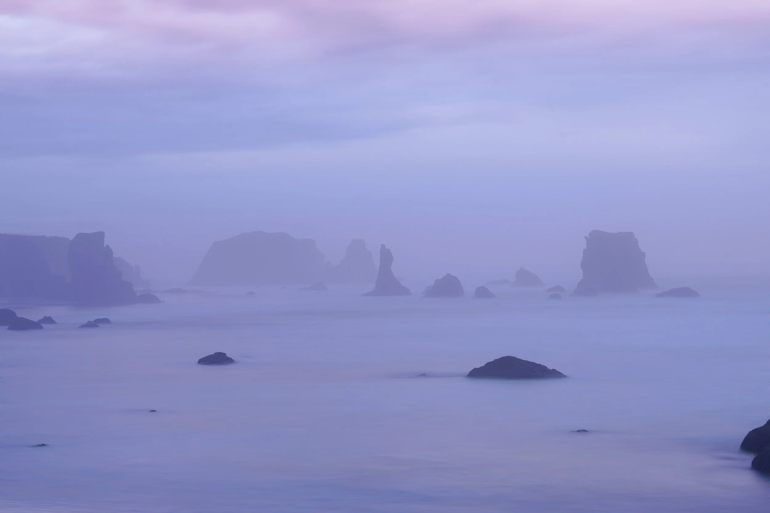 Foggy ocean scene with large rocks and formations sticking out of the water, under a cloudy sky.