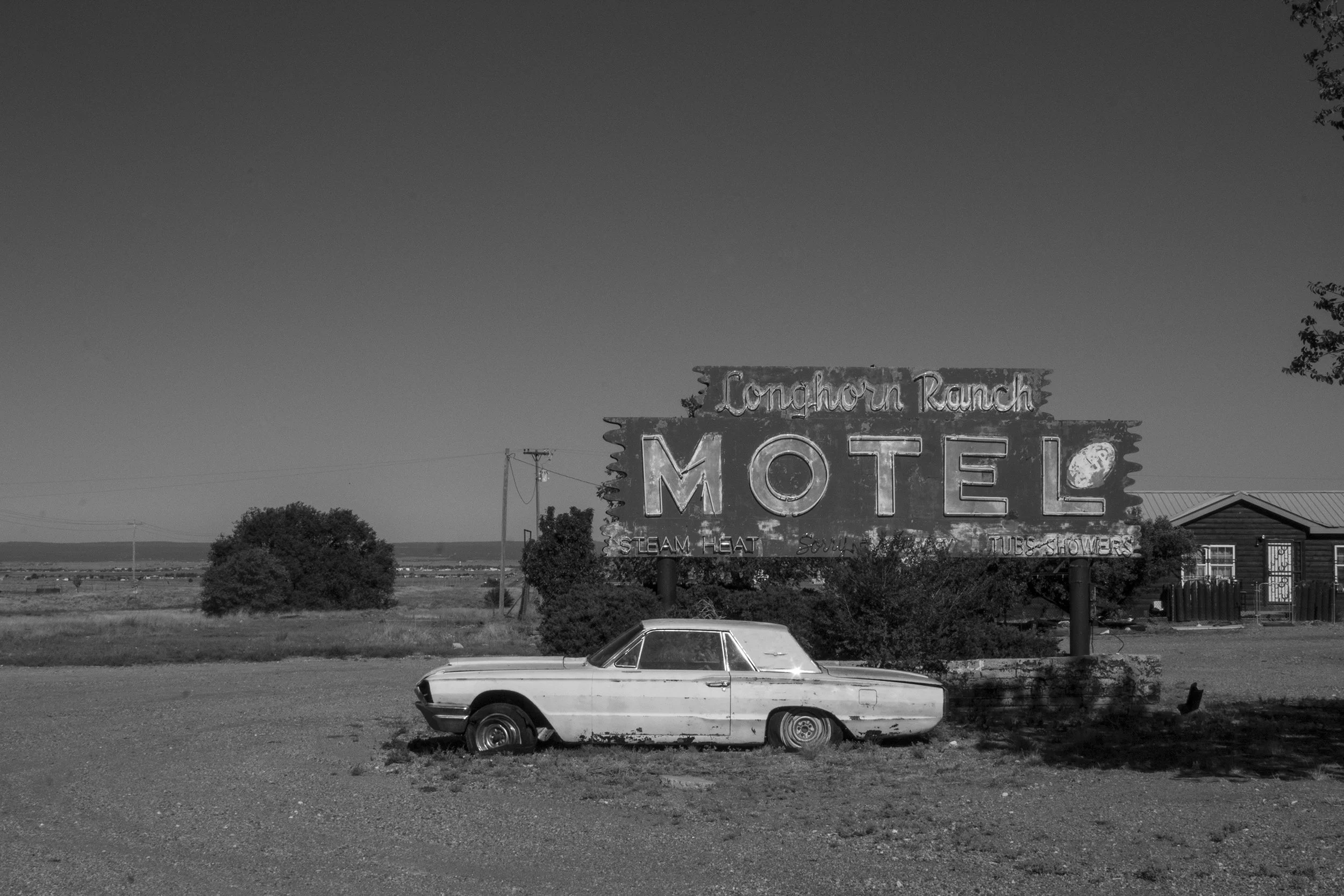 Black and white photo of a classic car parked on the grass in front of a sign that reads 'Longhorn Ranch Motel.' The sign also mentions features like steam heat, tubs, and showers. In the background, there are trees and a building, with a wide open l