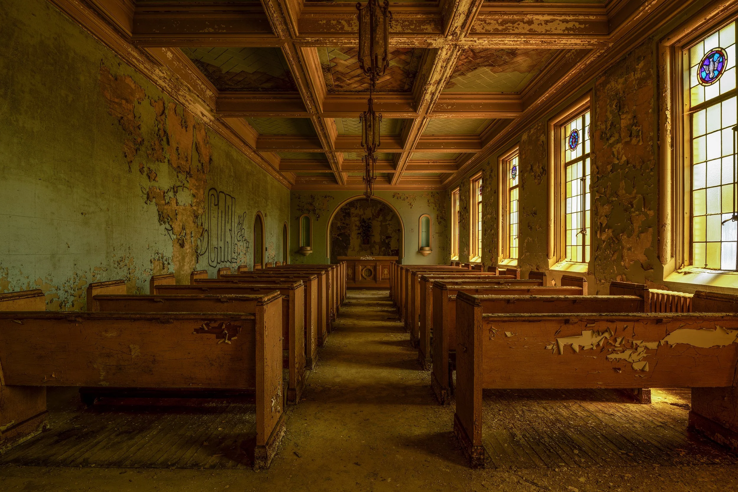 Interior of an abandoned church with peeling paint, broken pews, and stained glass windows.