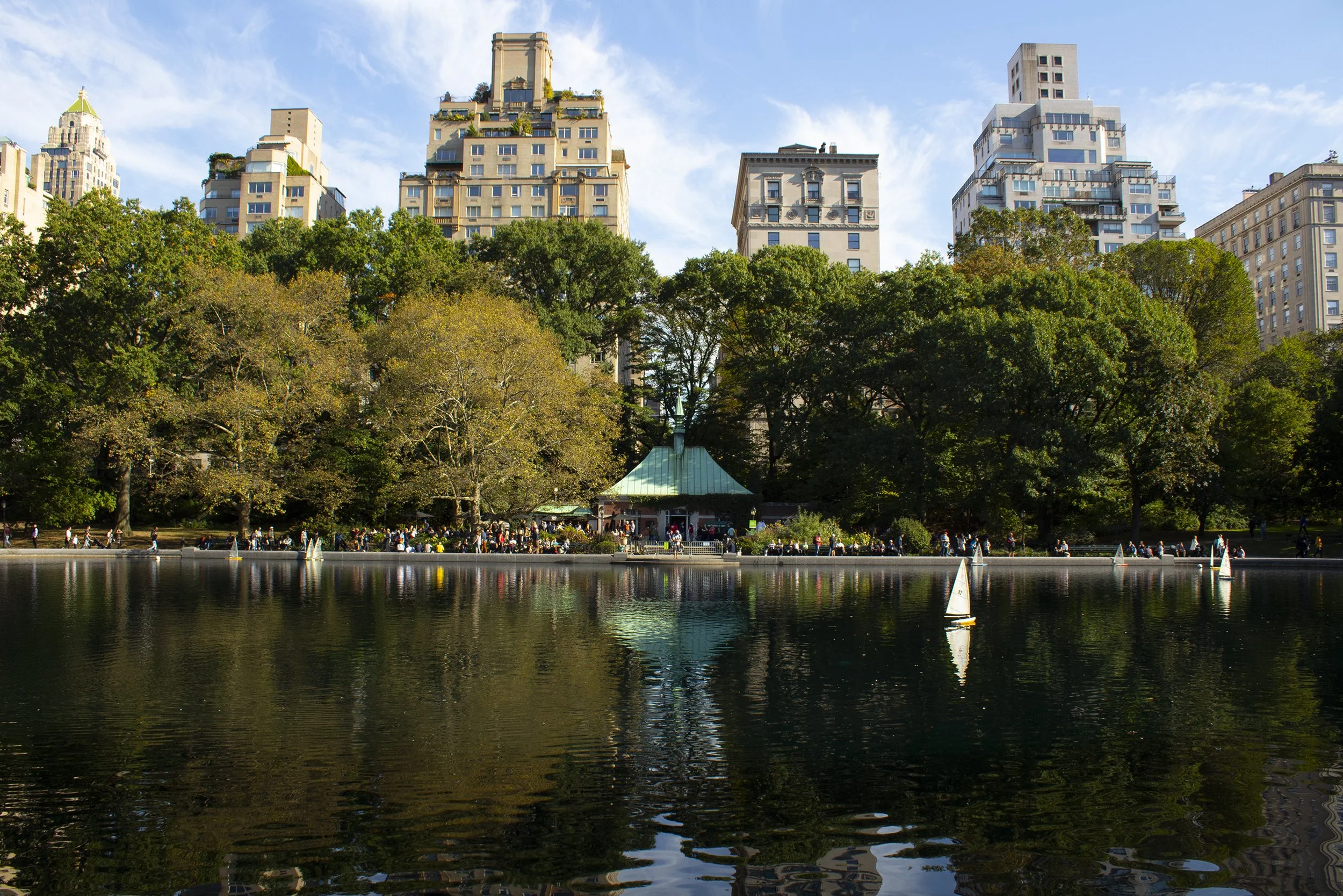 A park with trees and a pond with sailboats in the foreground, and tall buildings in the background under a partly cloudy sky.