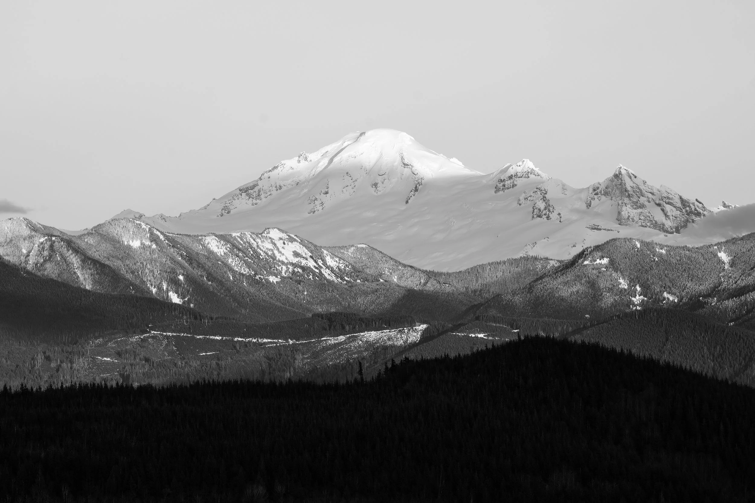 Snow-covered mountain rising above a forested landscape with a cloudy sky.